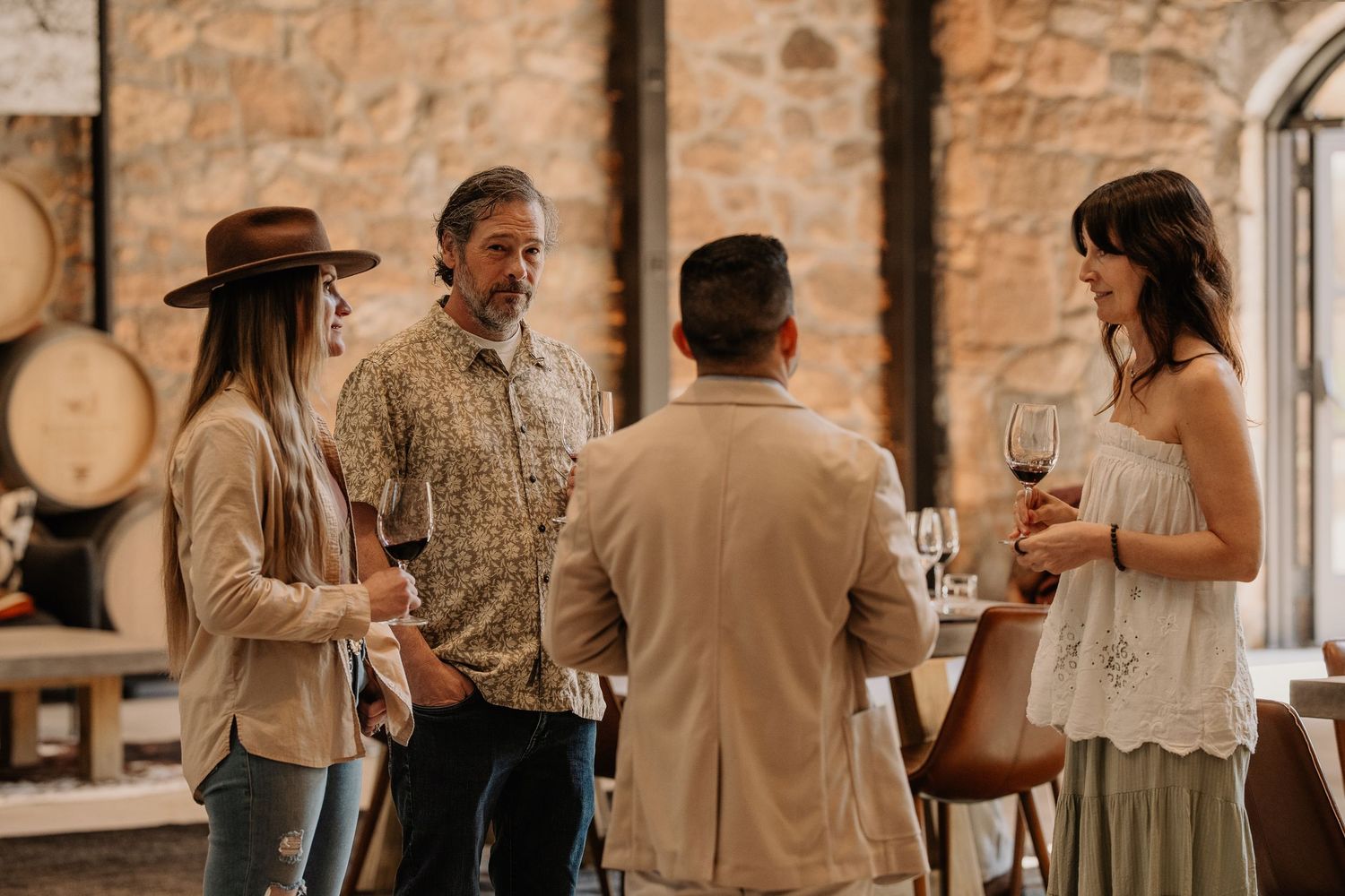Four adults socializing at a rustic winery, holding red wine glasses amid stone walls and oak barrels.