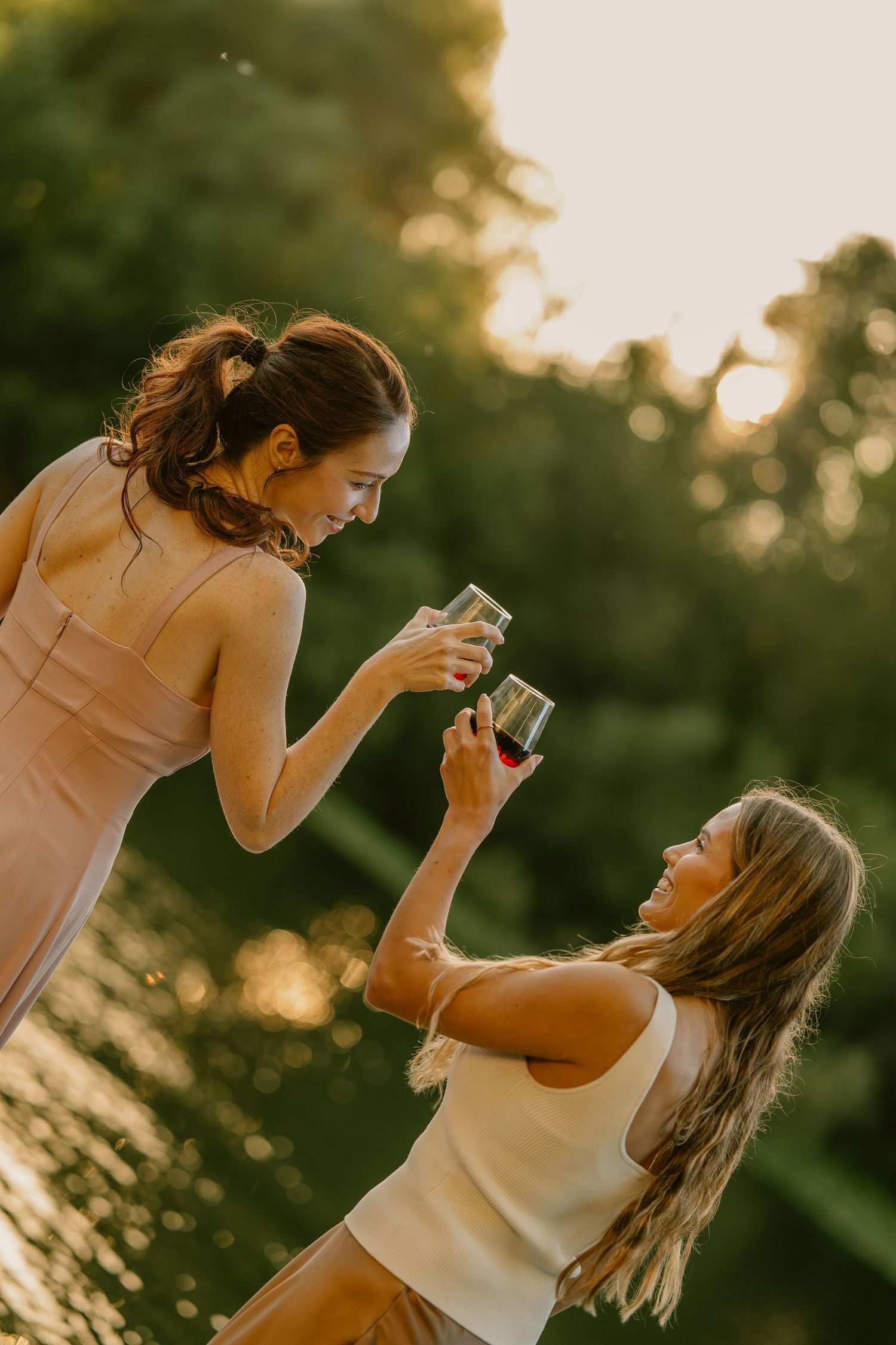 Two women toasting with wine glasses outdoors at golden hour, surrounded by lush green trees.