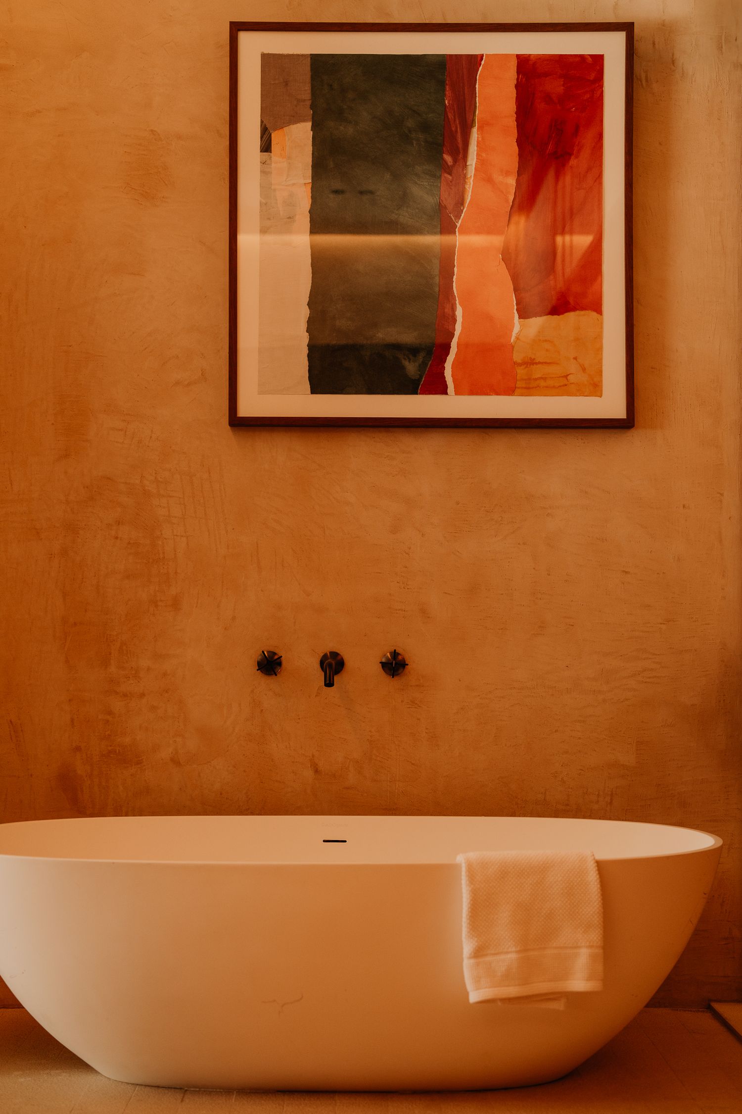 Modern white freestanding bathtub against terracotta wall with abstract art piece and black fixtures in luxury bathroom.