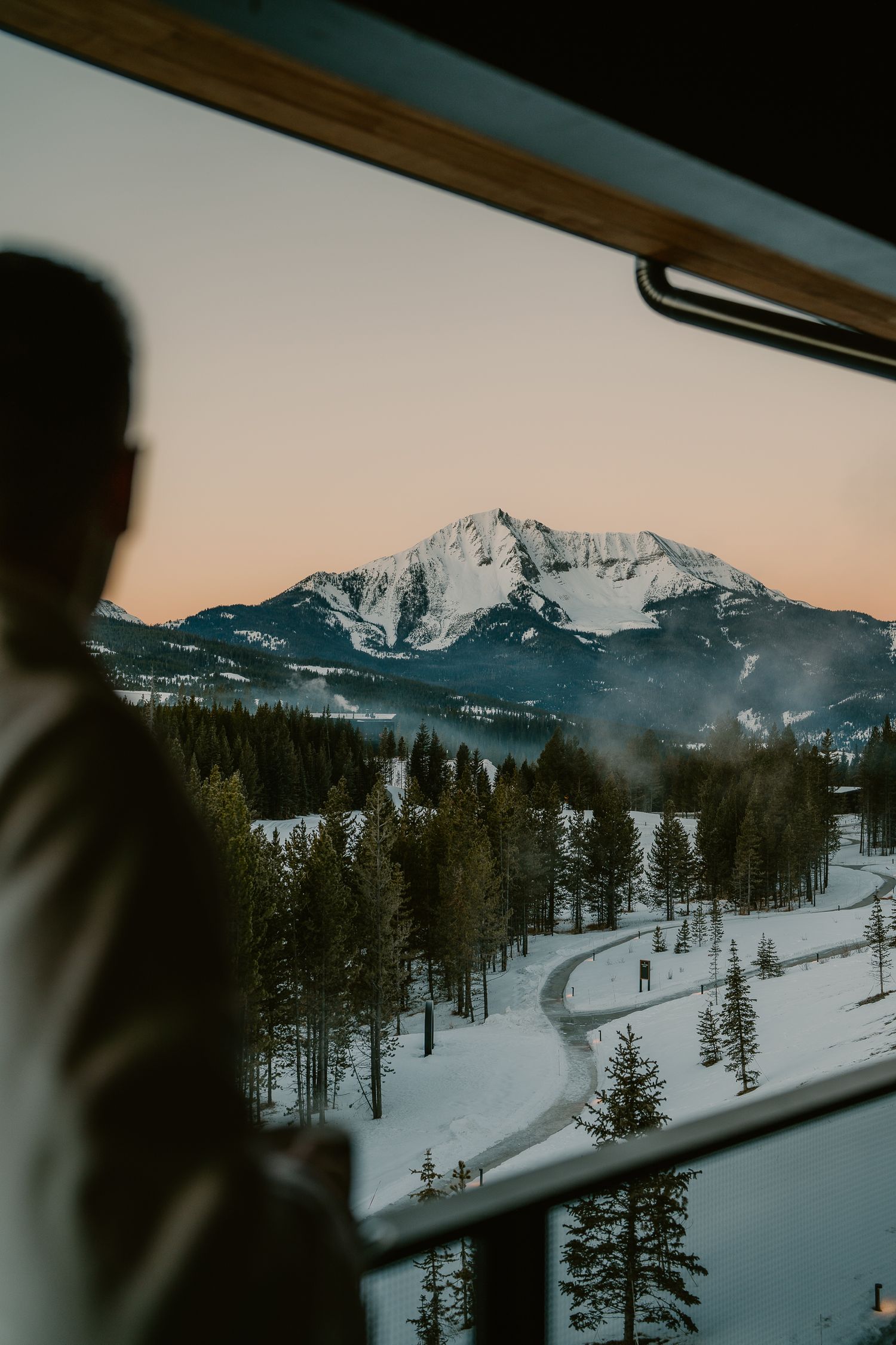 Snow-covered mountain peak and forest landscape viewed through a car window at sunset.