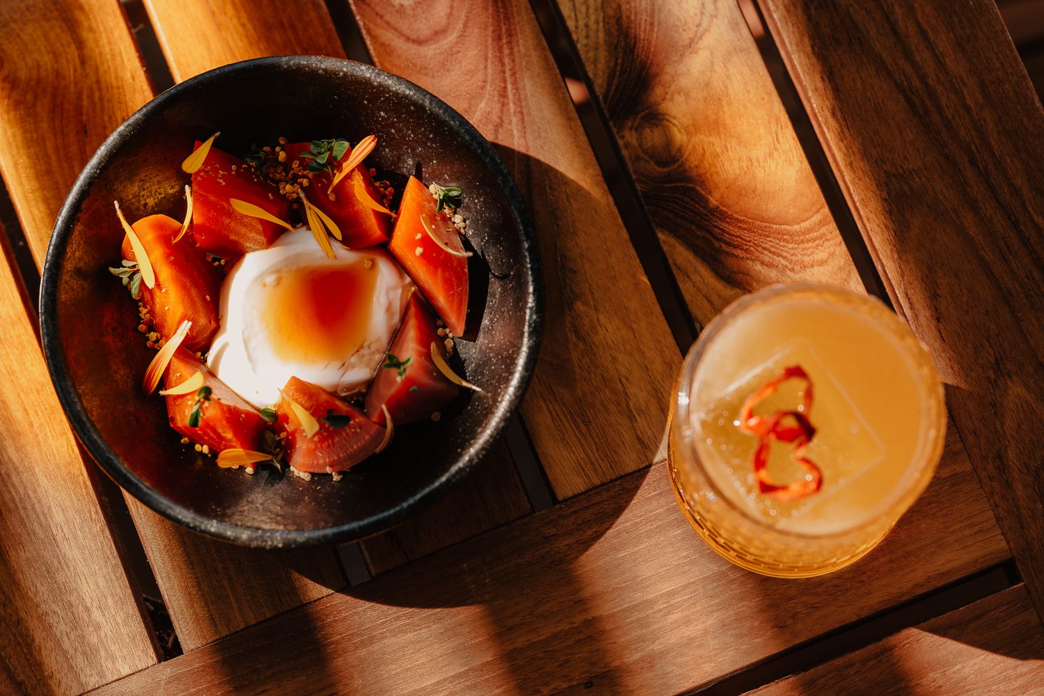 A poached egg bowl with smoked salmon and carrots beside a citrus cocktail on a wooden table.