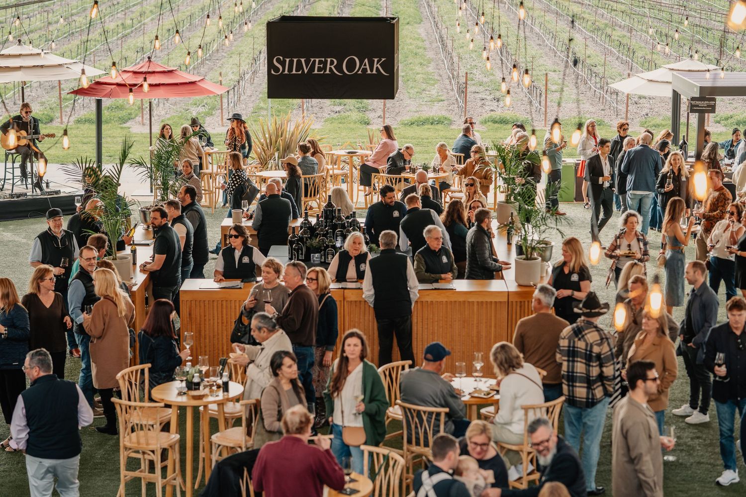 Crowded Silver Oak winery outdoor tasting event with guests mingling around wooden bar tables under string lights.