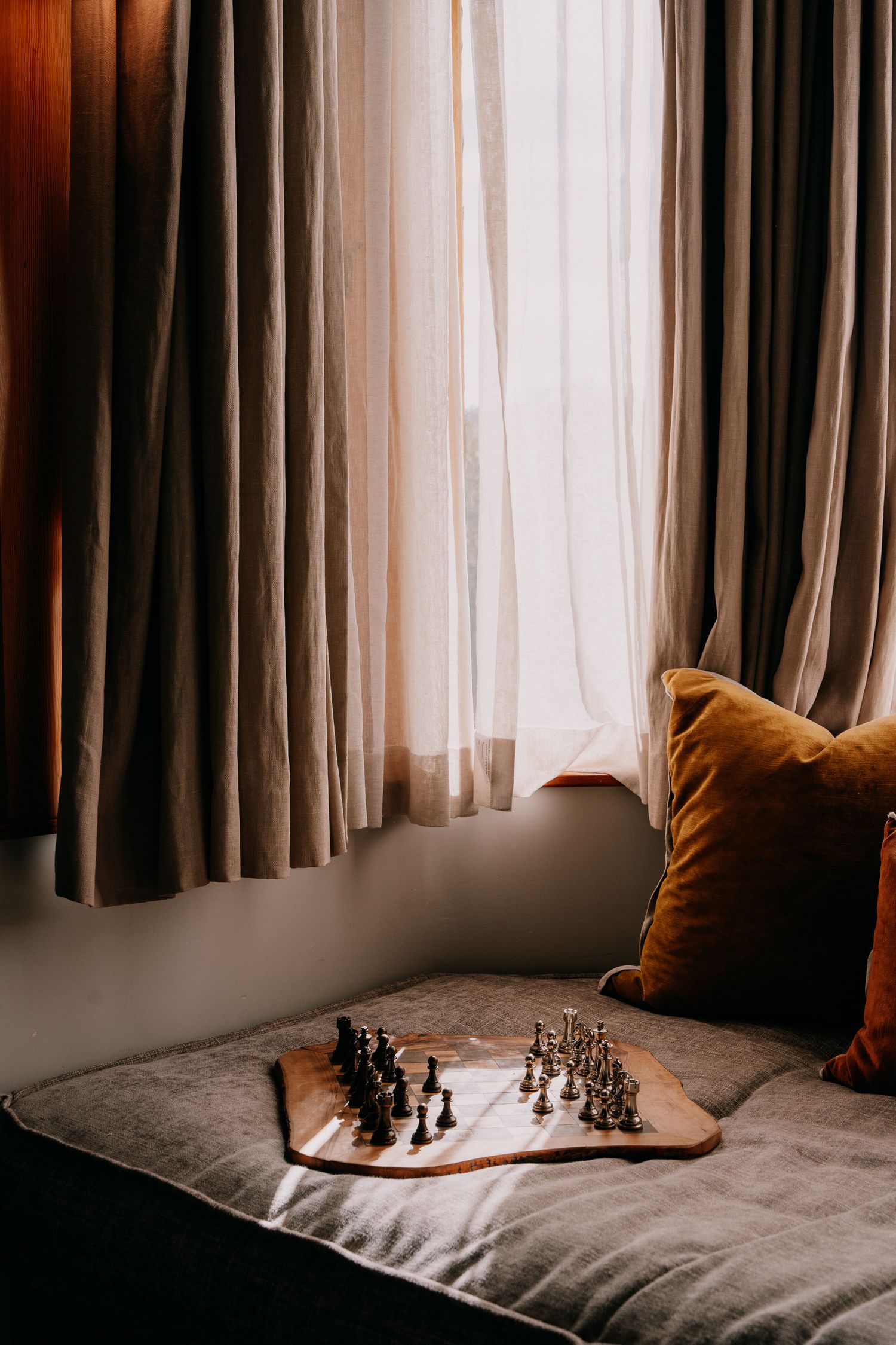 A chess set on a cozy windowsill with brown curtains and an orange velvet pillow.