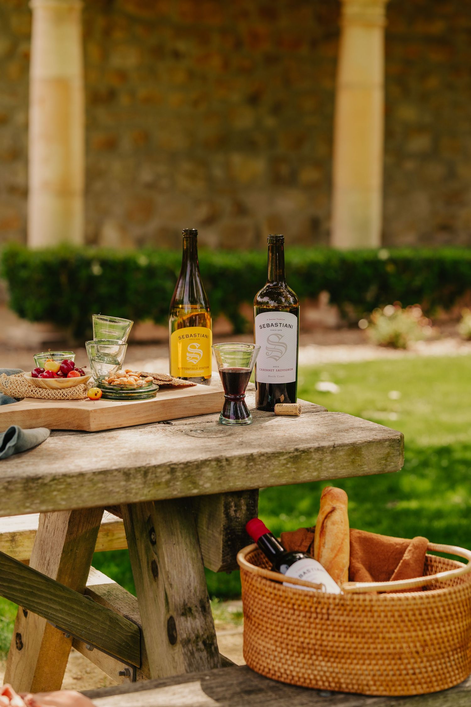 Sebastiani wine bottles on rustic wooden picnic table with charcuterie board and wicker basket outdoors.