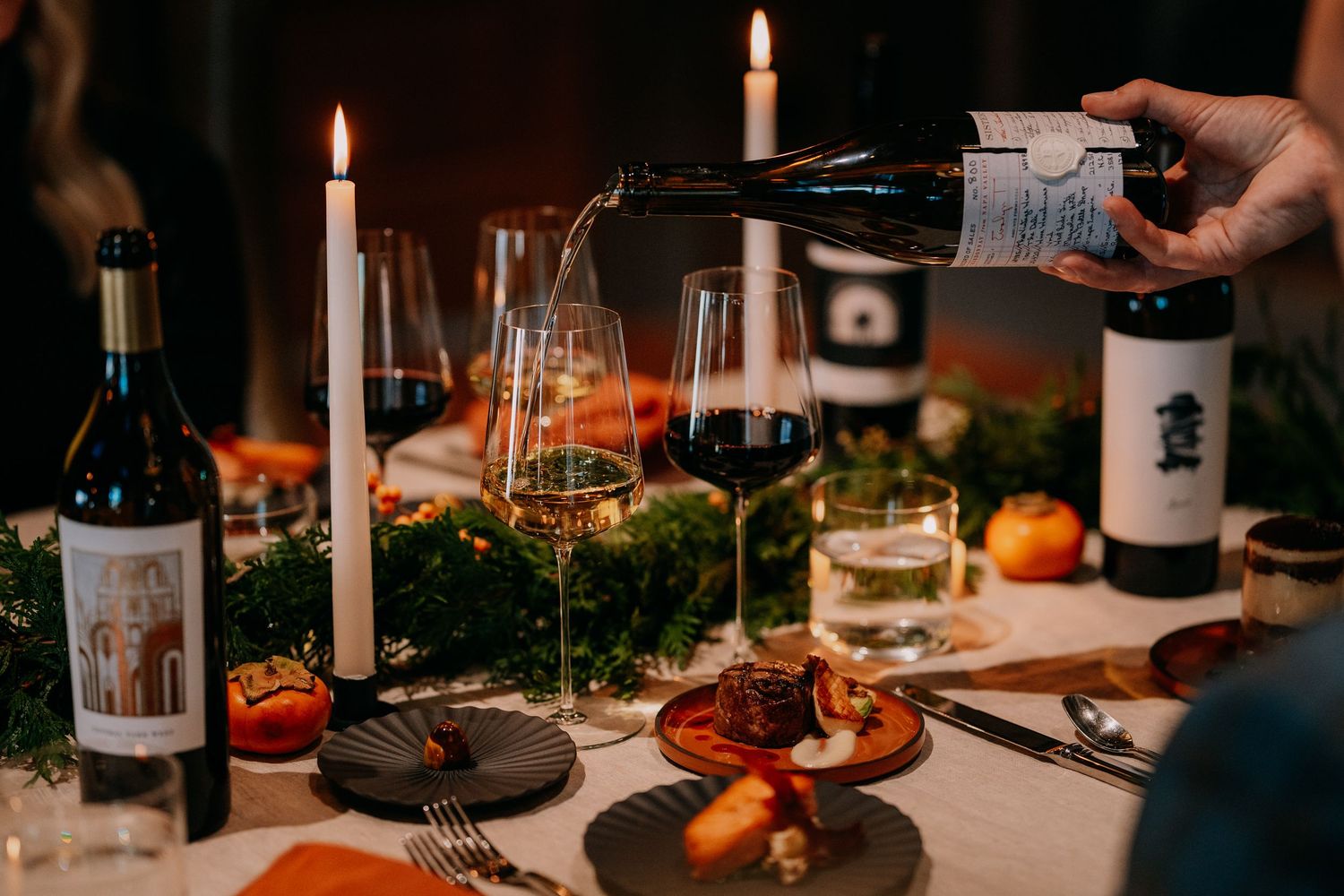 Red wine being poured at an intimate candlelit dinner table with seasonal decorations and fresh citrus fruits.