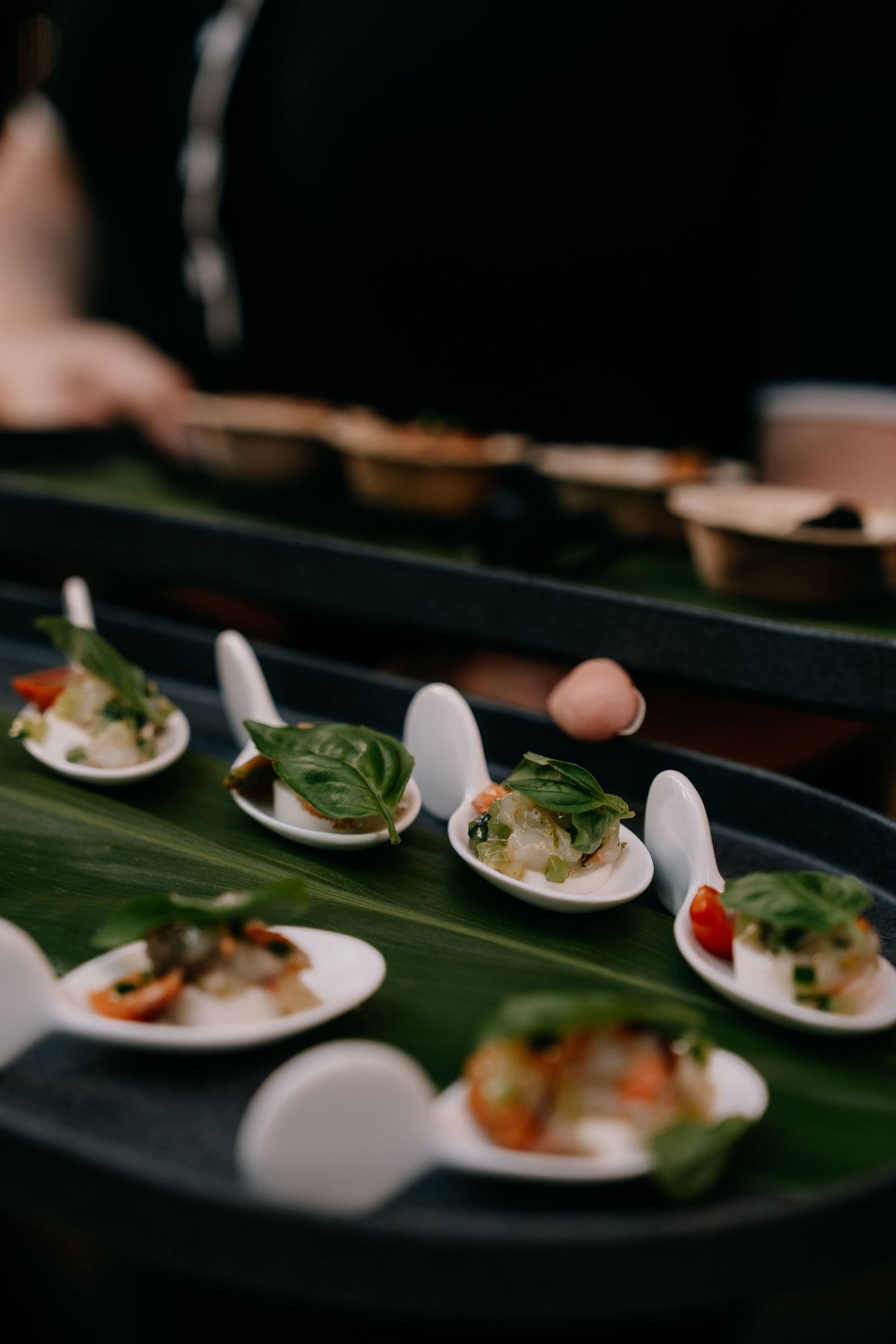 Elegant canapés served on white ceramic spoons arranged on dark green banana leaves in a fine dining setting.