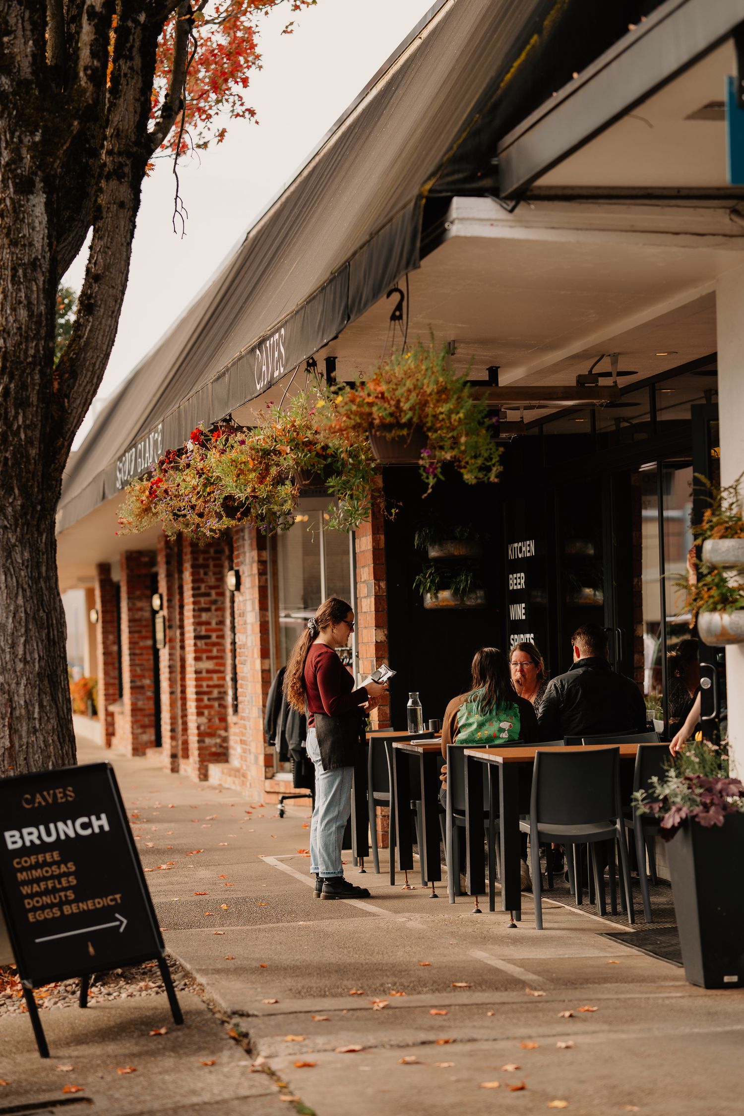 People enjoying outdoor dining at a cozy brunch spot with hanging flower baskets and brick storefront on an autumn day.