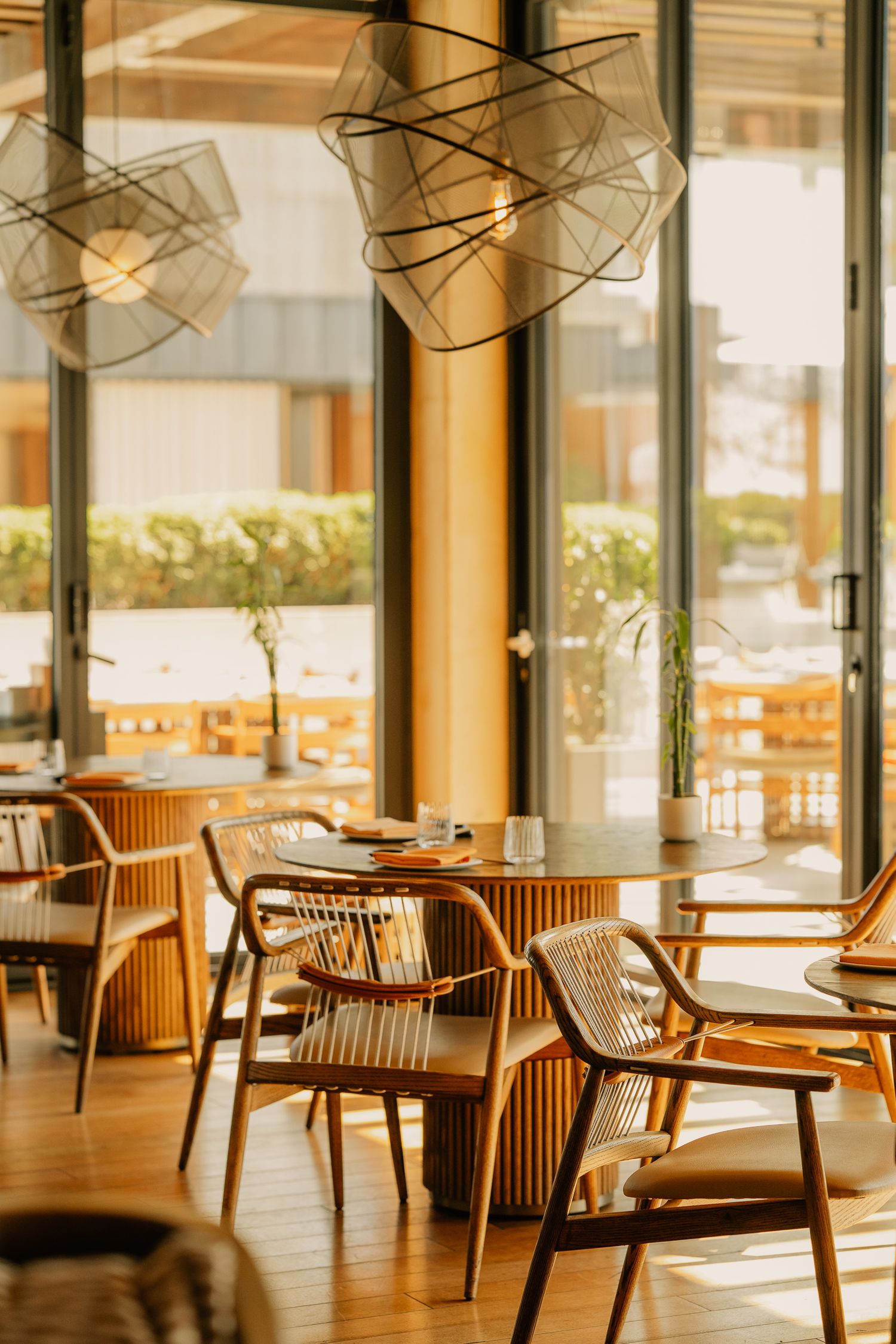 Bright modern restaurant interior with wooden chairs, round tables, geometric pendant lights, and floor-to-ceiling windows.