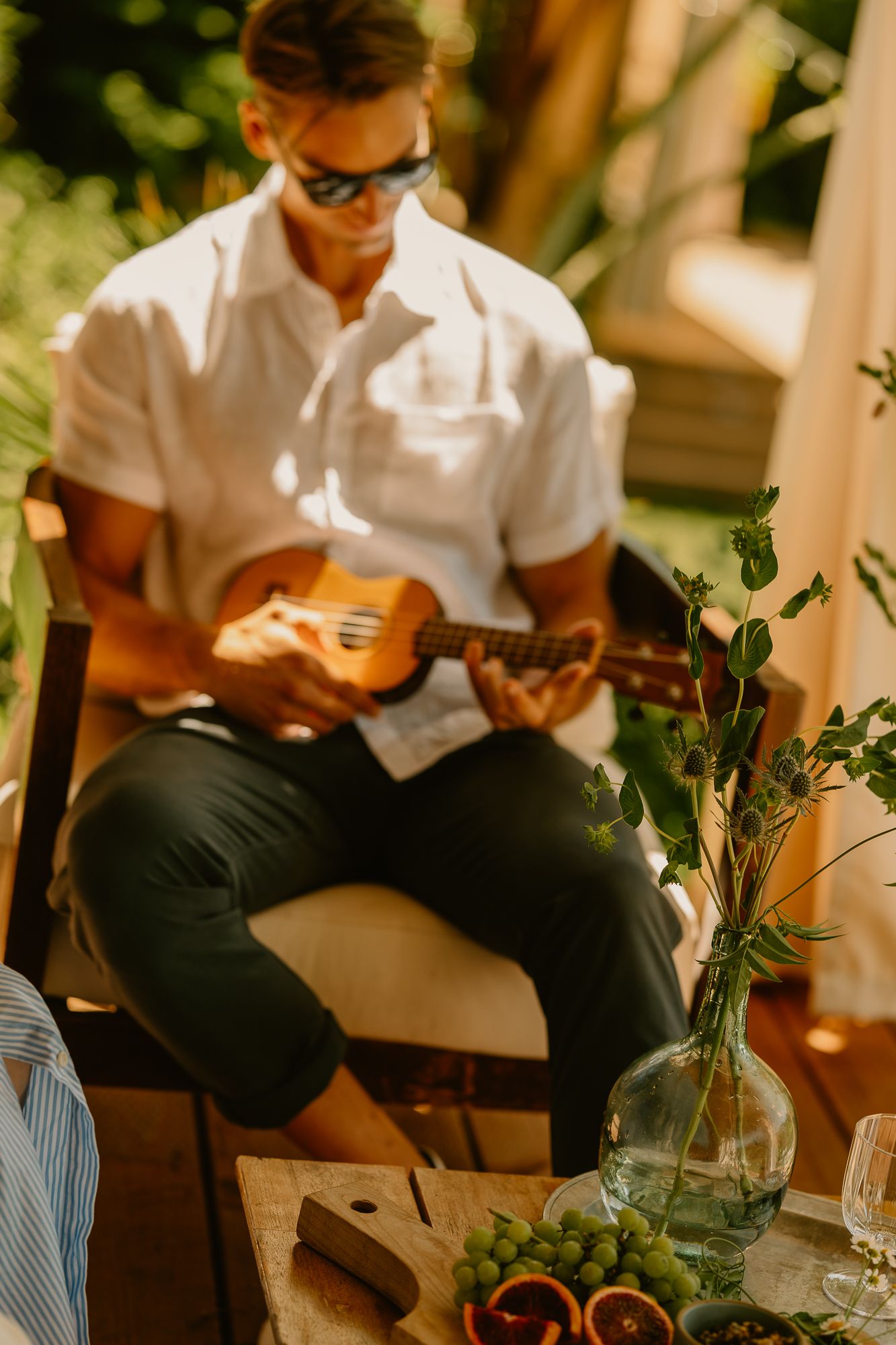 Man in white shirt playing ukulele outdoors at a sunlit table with grapes, citrus, and a glass vase with greenery.