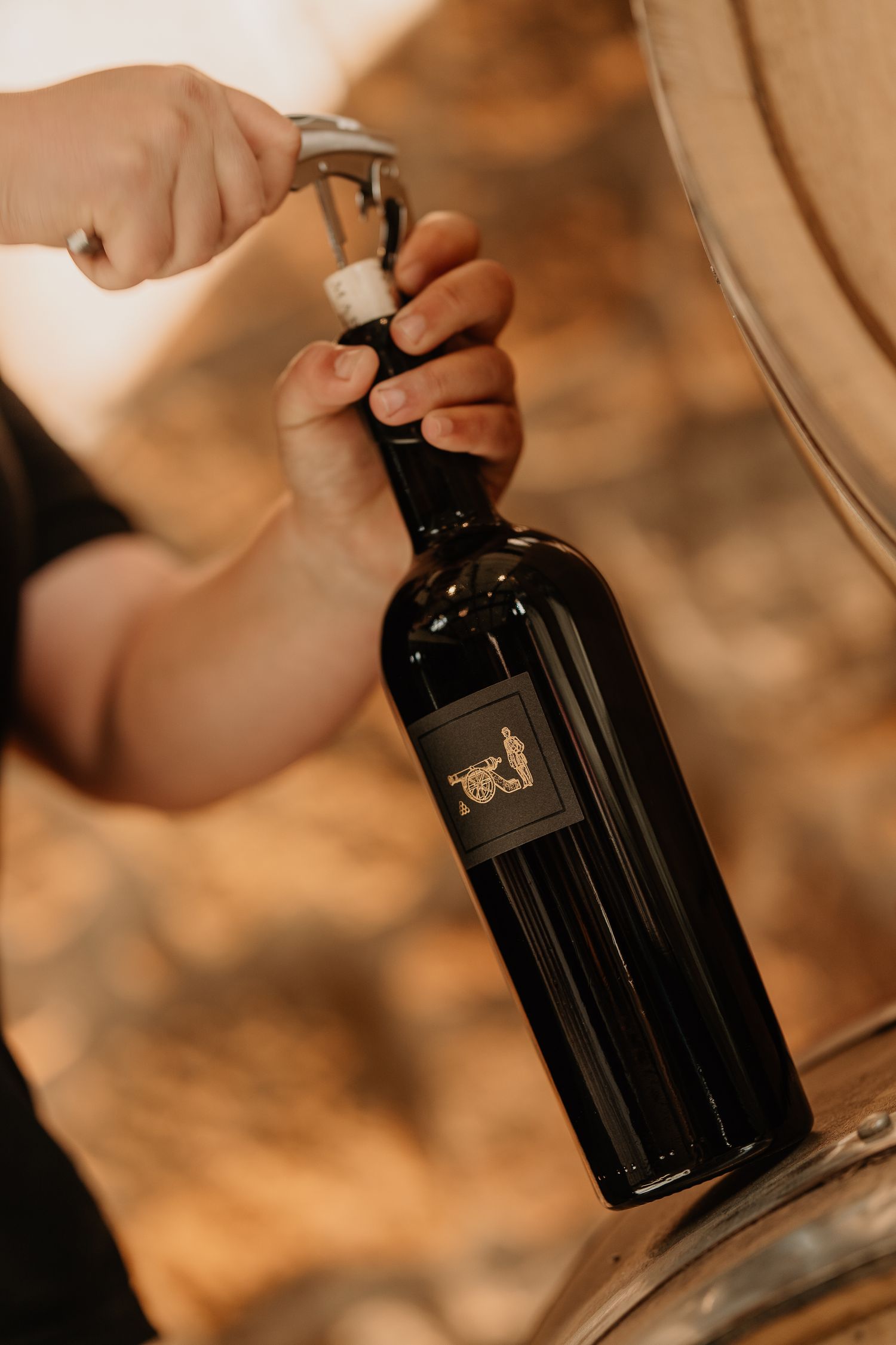 Hands filling a dark wine bottle from an oak barrel using a wine thief tool.