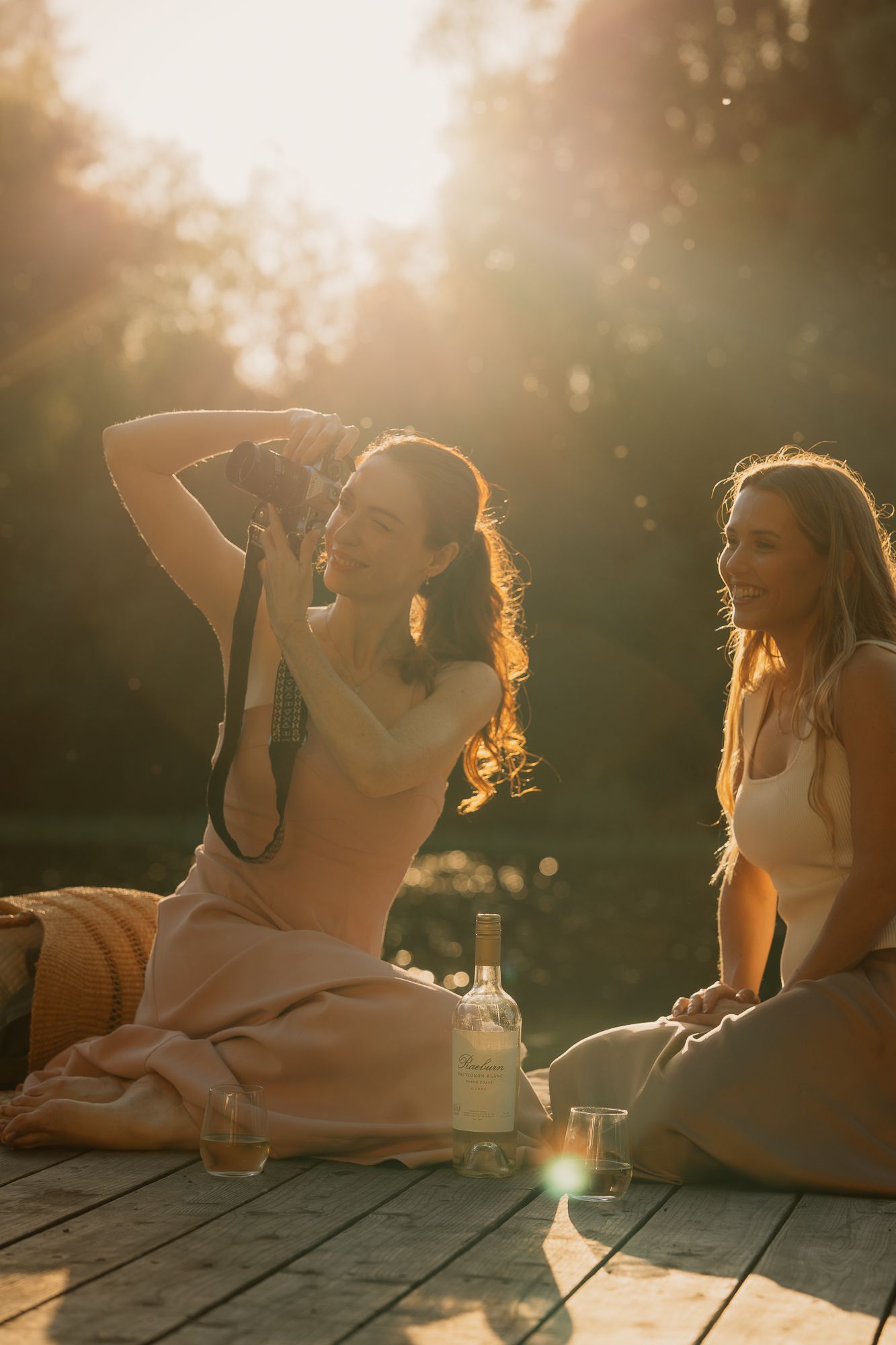 Two women sitting on a wooden deck in golden sunlight, laughing and relaxing with a bottle of wine.