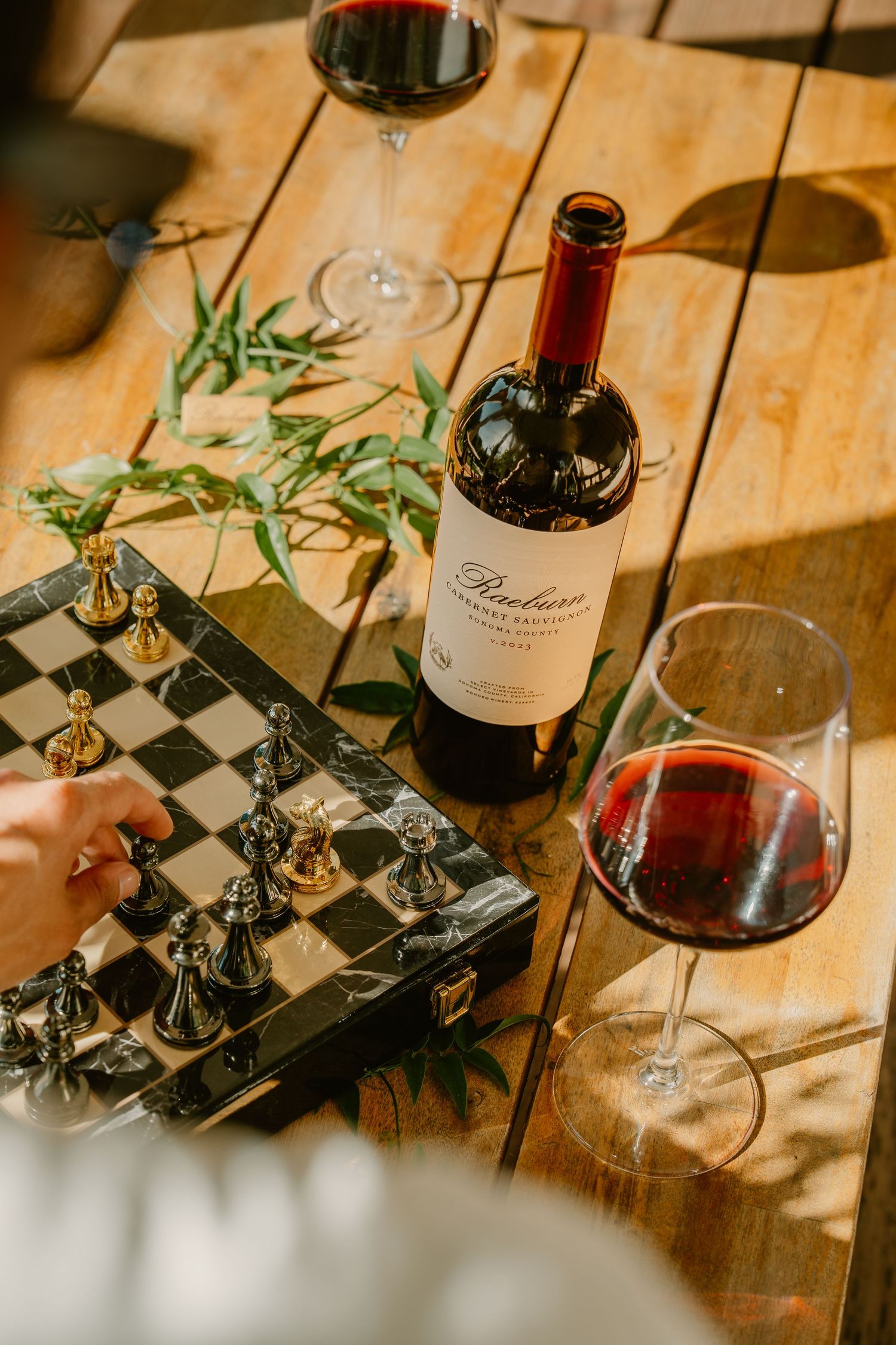 A bottle of red wine beside a filled glass and a chess board on a wooden table in warm sunlight.