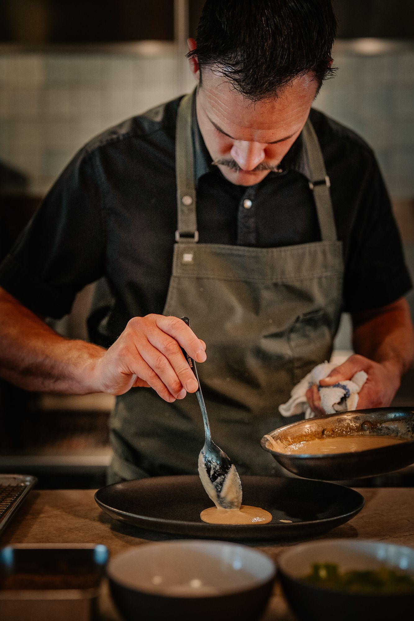 Professional chef plating a gourmet dish with a spoon in a modern restaurant kitchen.