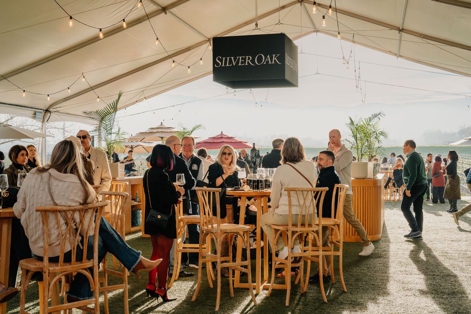 Guests sit and socialize under a Silver Oak branded tent at an outdoor winery event on a sunny day.