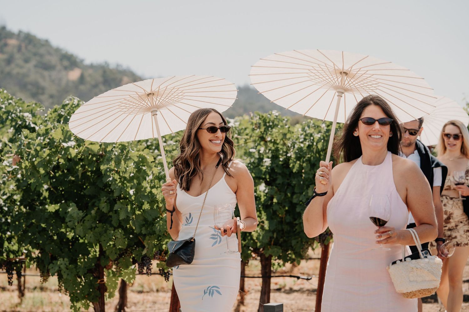 Two women holding white parasol umbrellas walking through sunny vineyard rows at outdoor wine event.