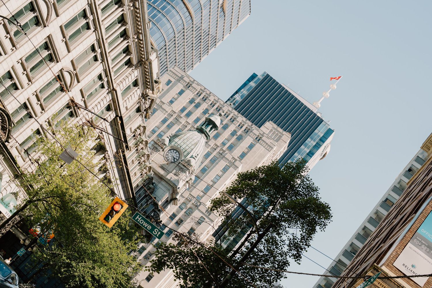 Looking up at modern skyscrapers and classical architecture in New York City with trees in the foreground against blue sky.