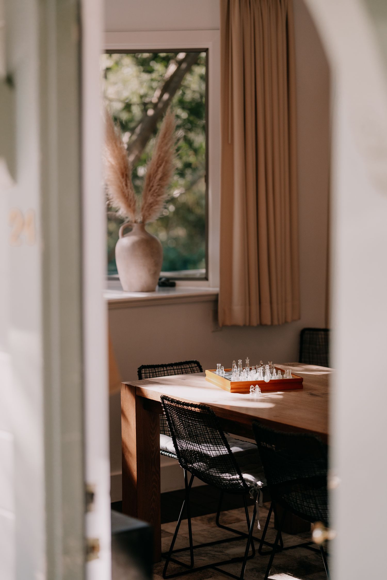Cozy dining area with wooden table, folding chairs, chess set and pampas grass in vase by window with tan curtains.