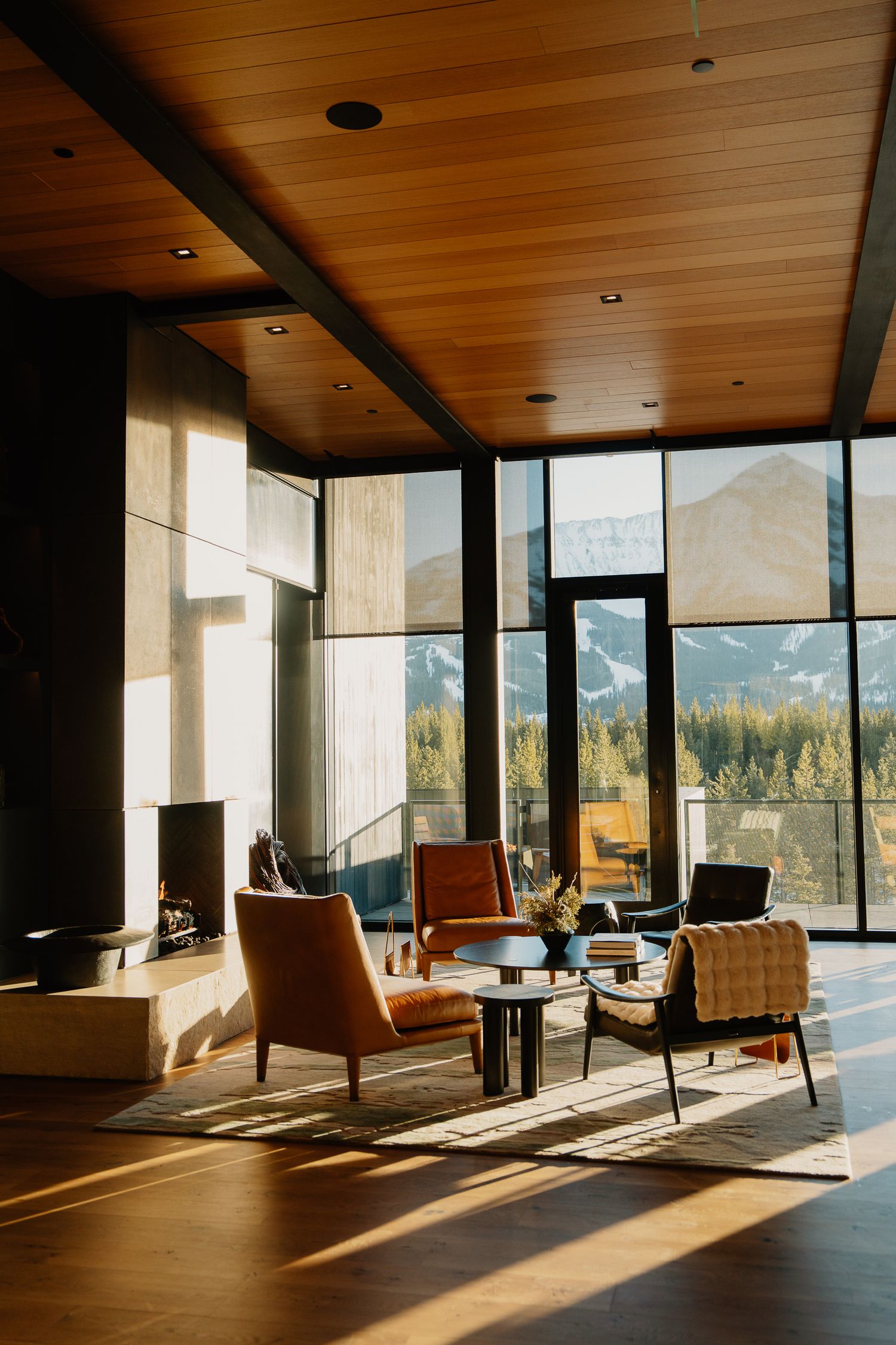 Modern mountain lodge interior with floor-to-ceiling windows, wooden ceiling, and mid-century modern furniture bathed in sunlight.