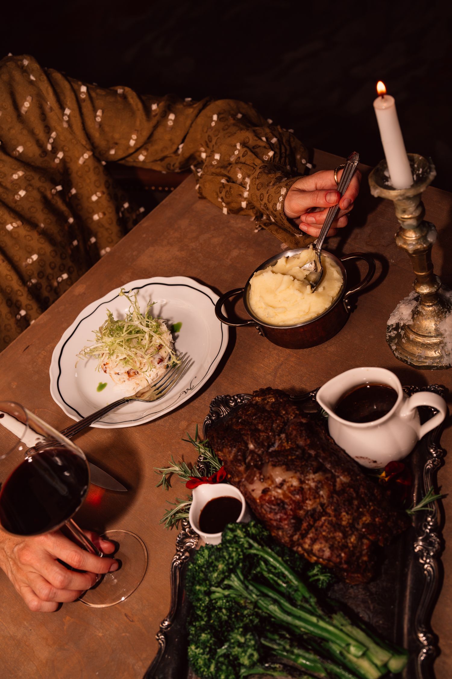 Elegant dinner setting with broccolini, mashed potatoes and meat dish served on white plates in dim lighting.