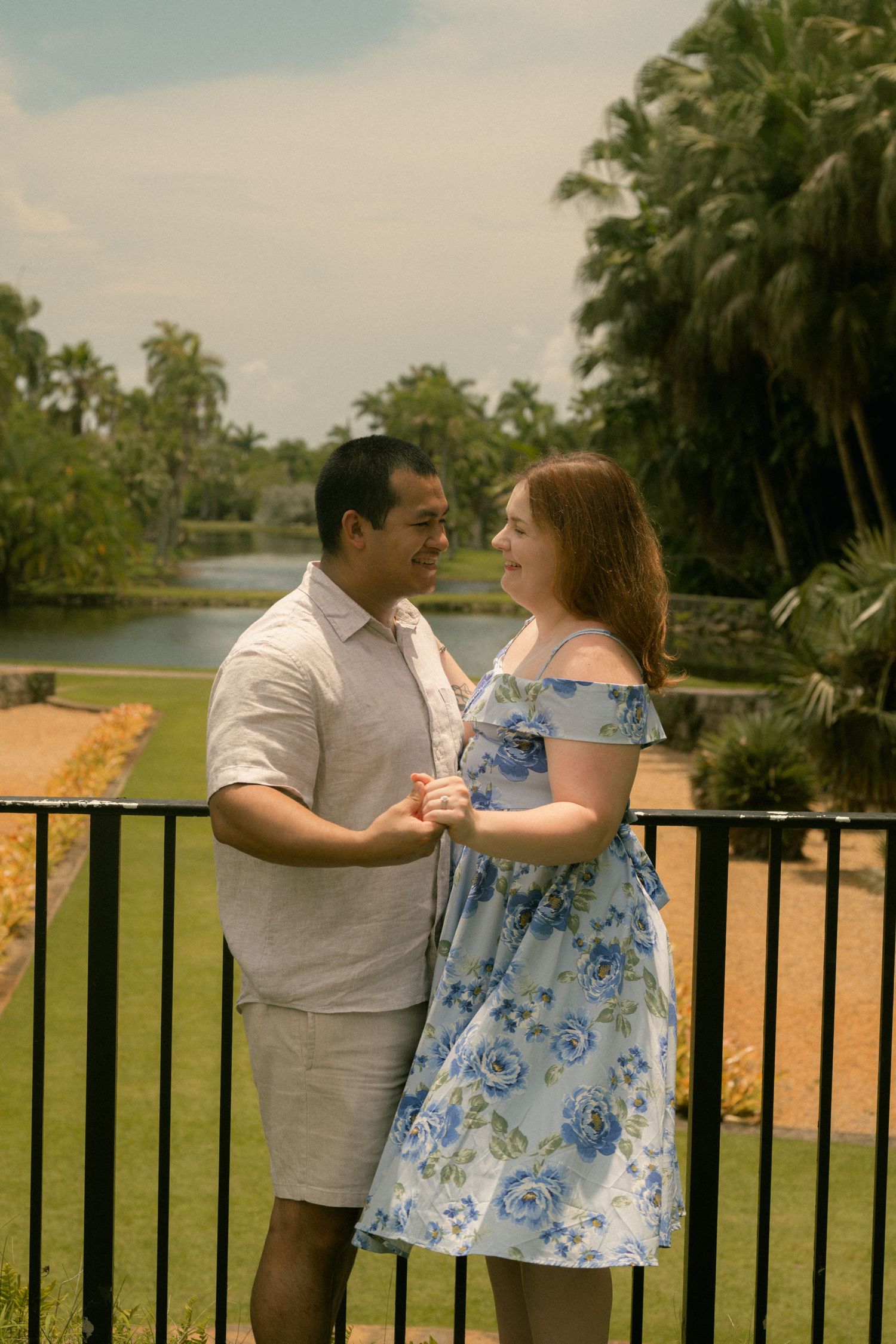 Couple holding hands and gazing at each other on a balcony surrounded by lush tropical gardens and a pond.