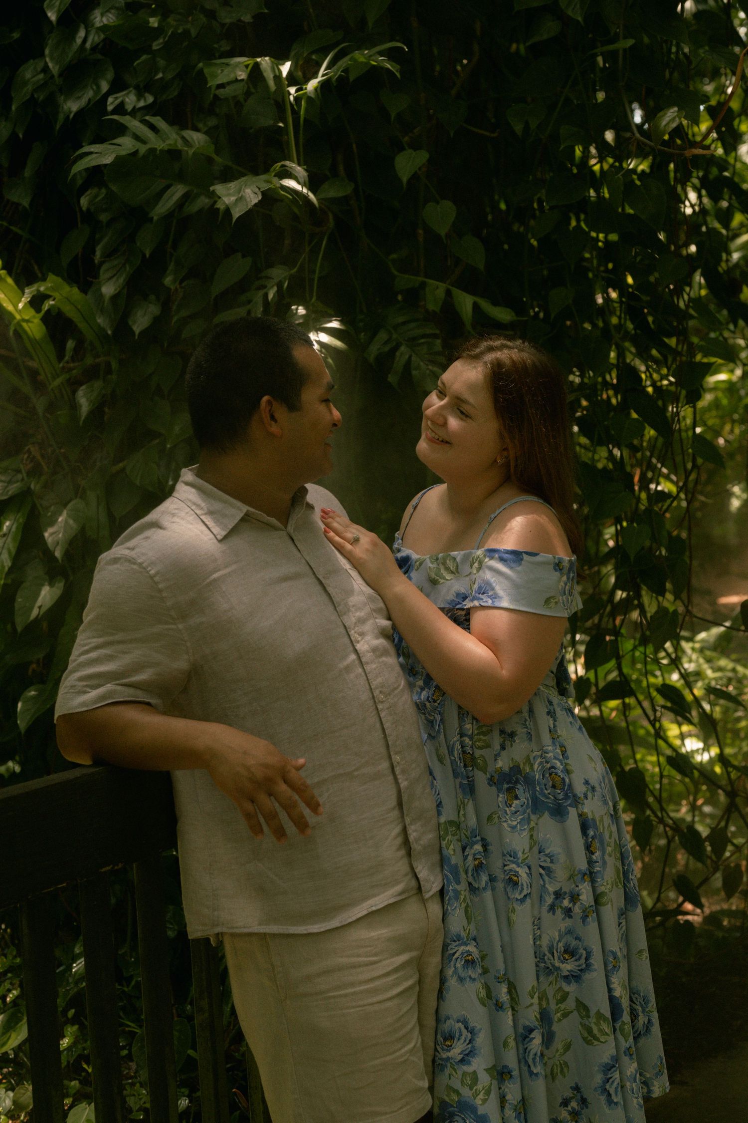 Couple sharing a romantic moment surrounded by lush tropical greenery, woman in blue floral dress touching man's chest.