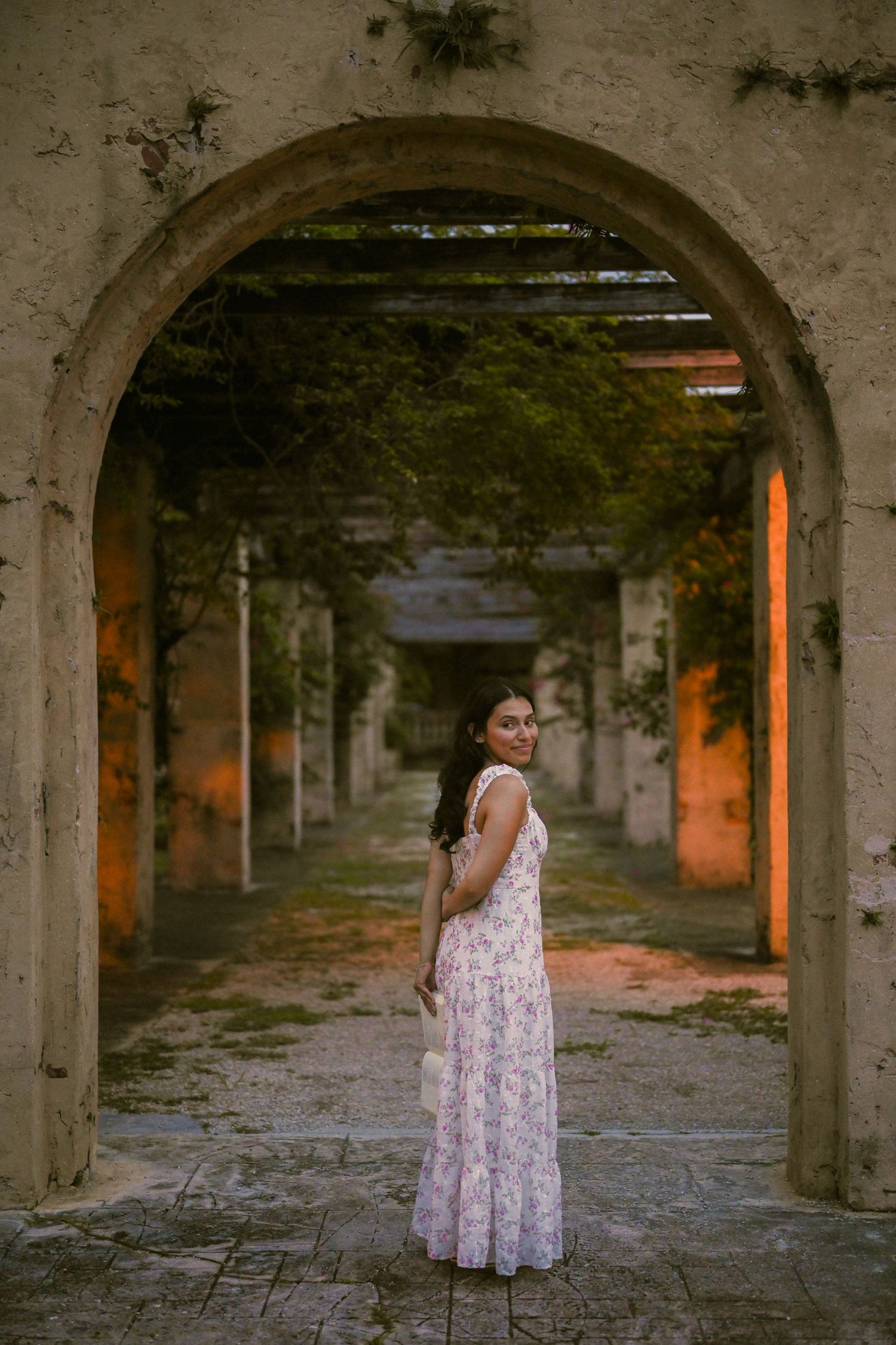 A person in a floral dress stands under stone archways in a rustic setting.