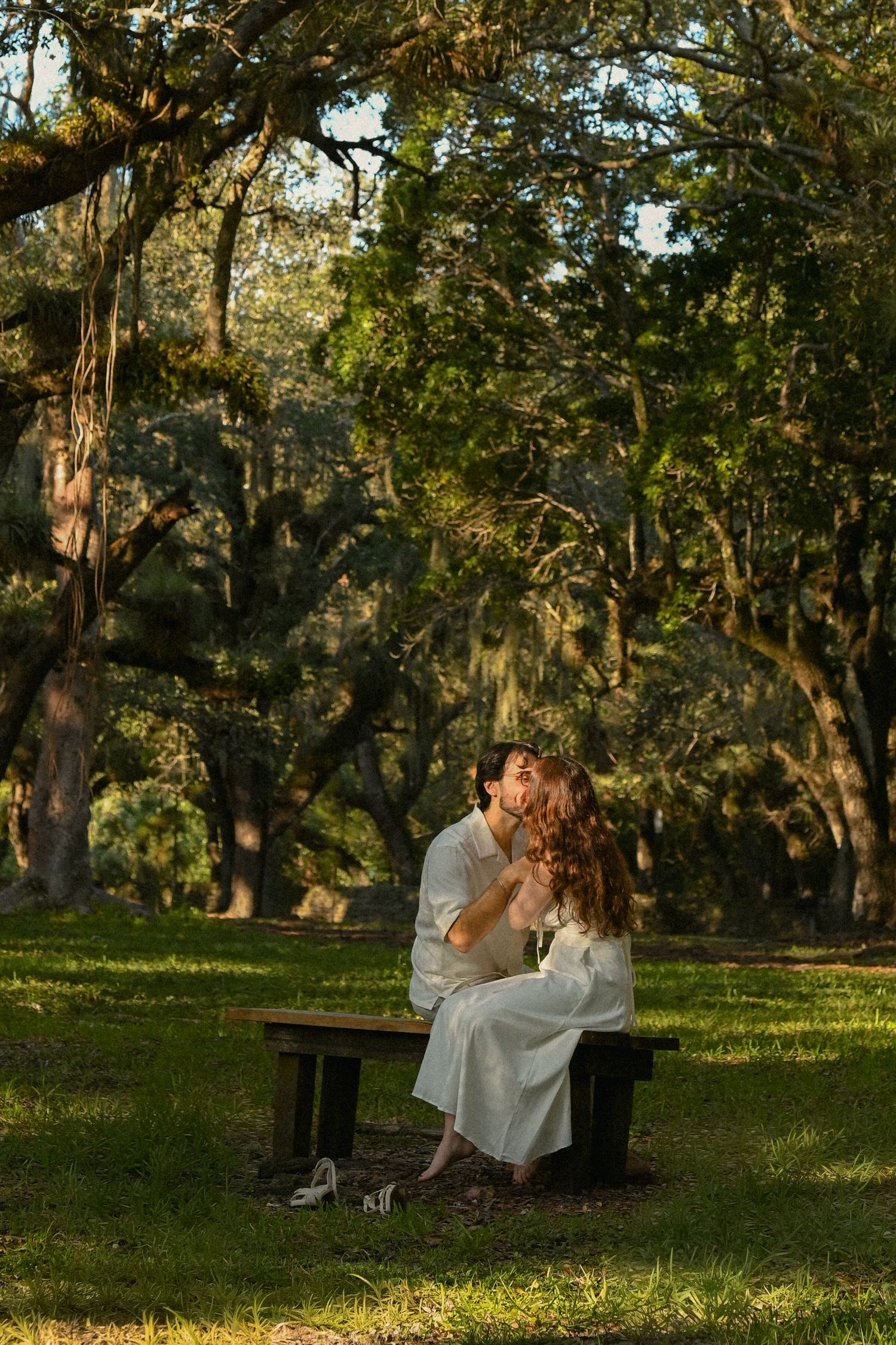 A couple sits together on a wooden bench in a sunlit park surrounded by moss-draped oak trees.