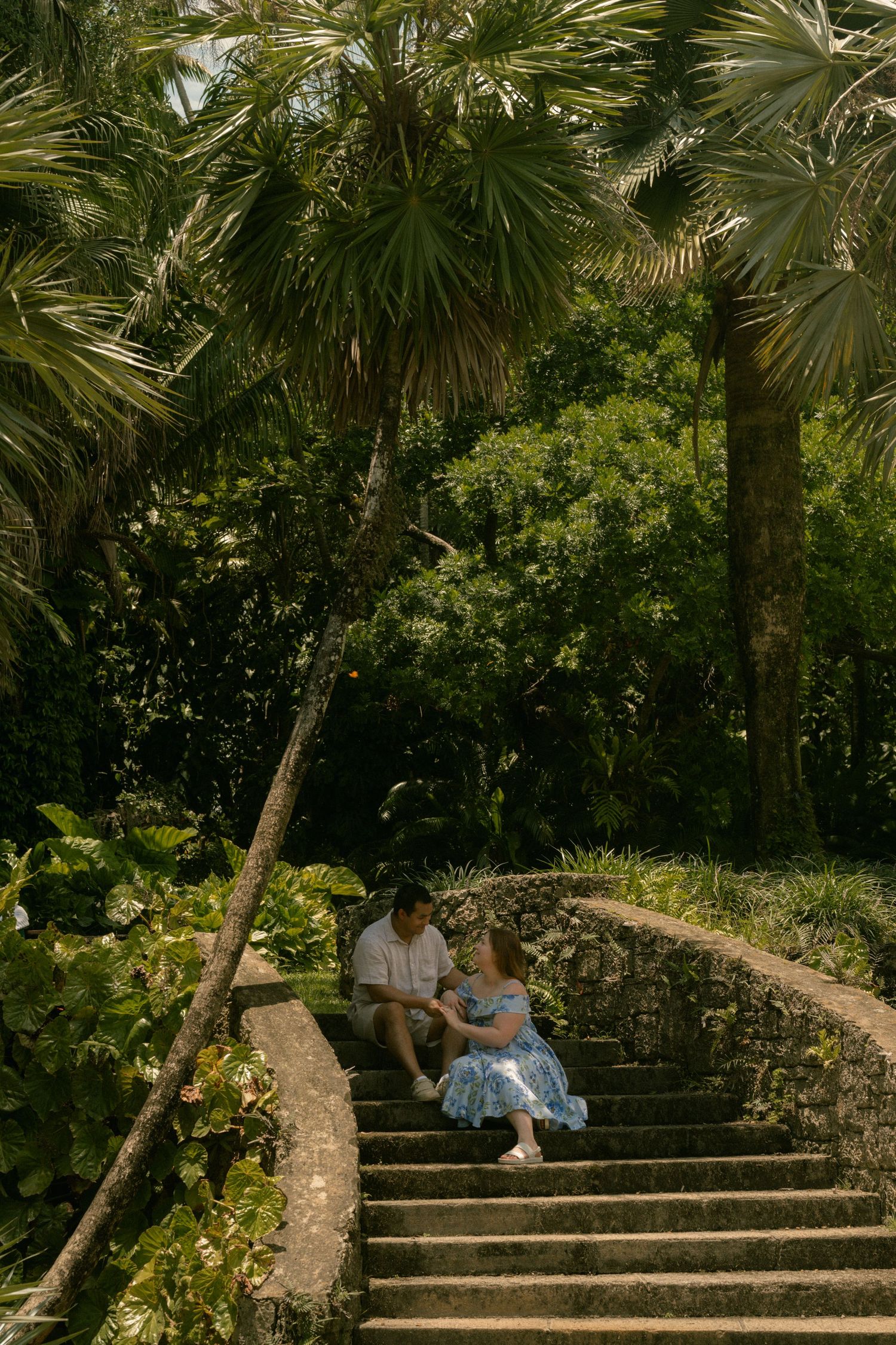 Couple sitting on stone steps surrounded by lush tropical palm trees and greenery in a serene garden setting.