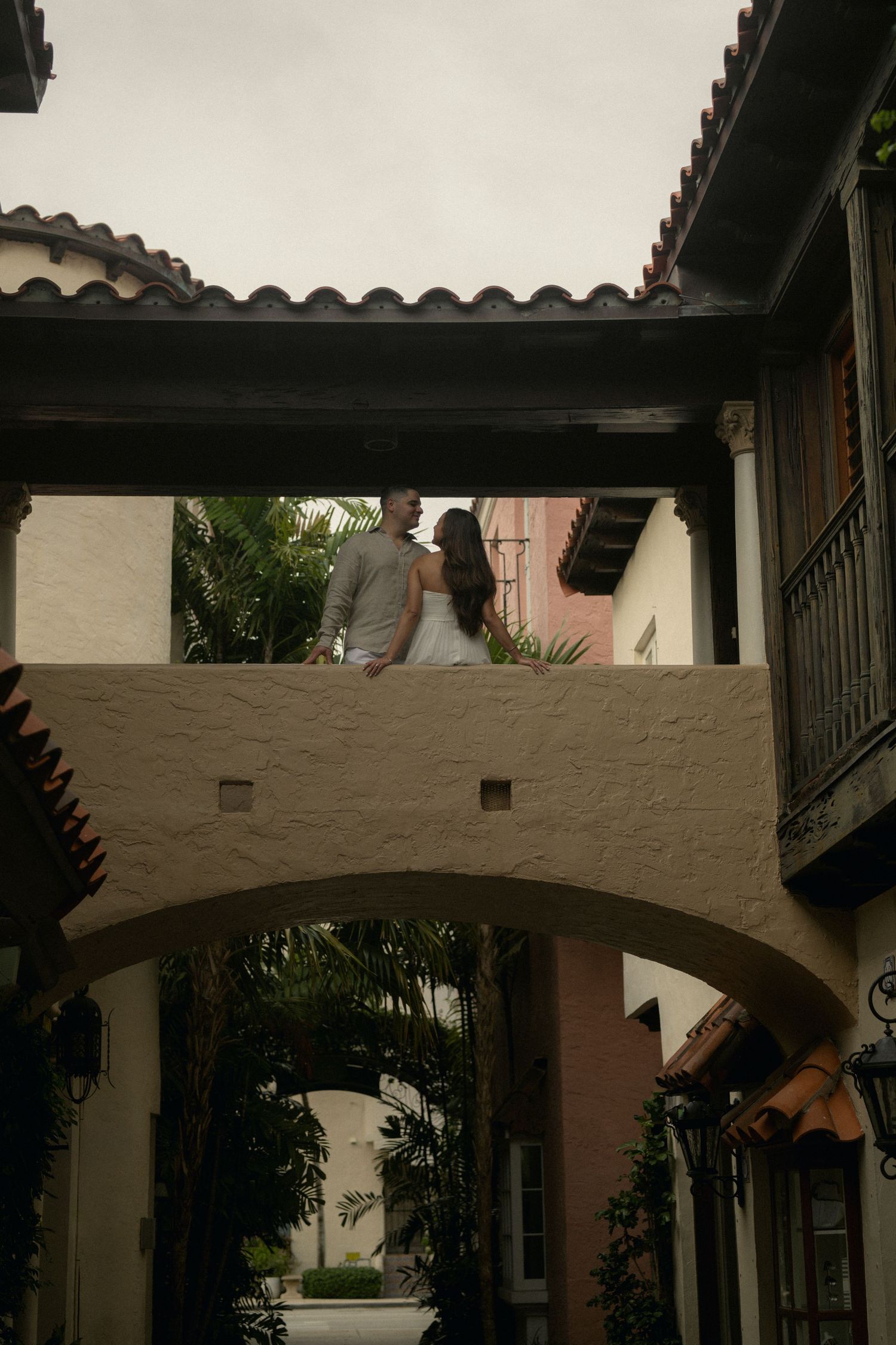 Couple holding hands on a Spanish-style archway bridge surrounded by lush tropical greenery.