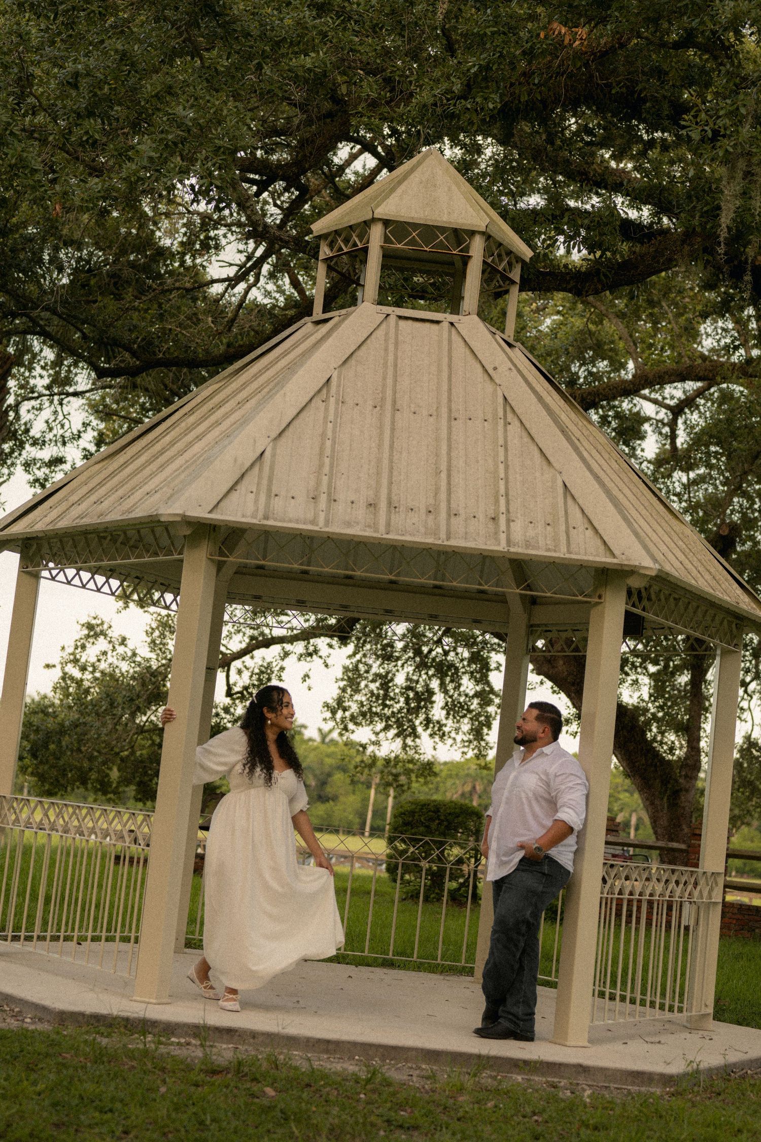 Couple in white outfits dancing under a wooden gazebo surrounded by lush green trees in a park.