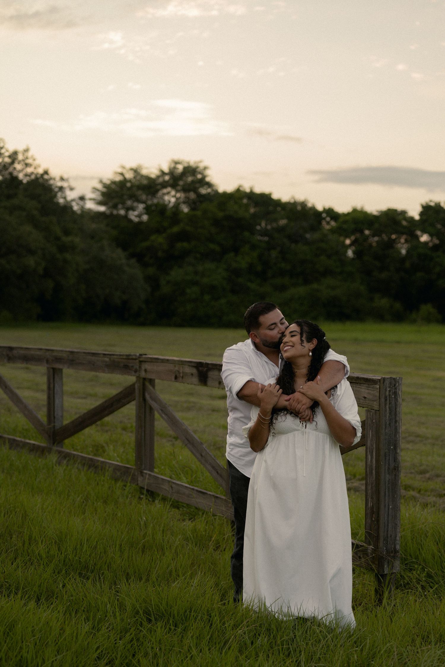 Man in white shirt hugging woman in white dress by wooden fence in green meadow at dusk.
