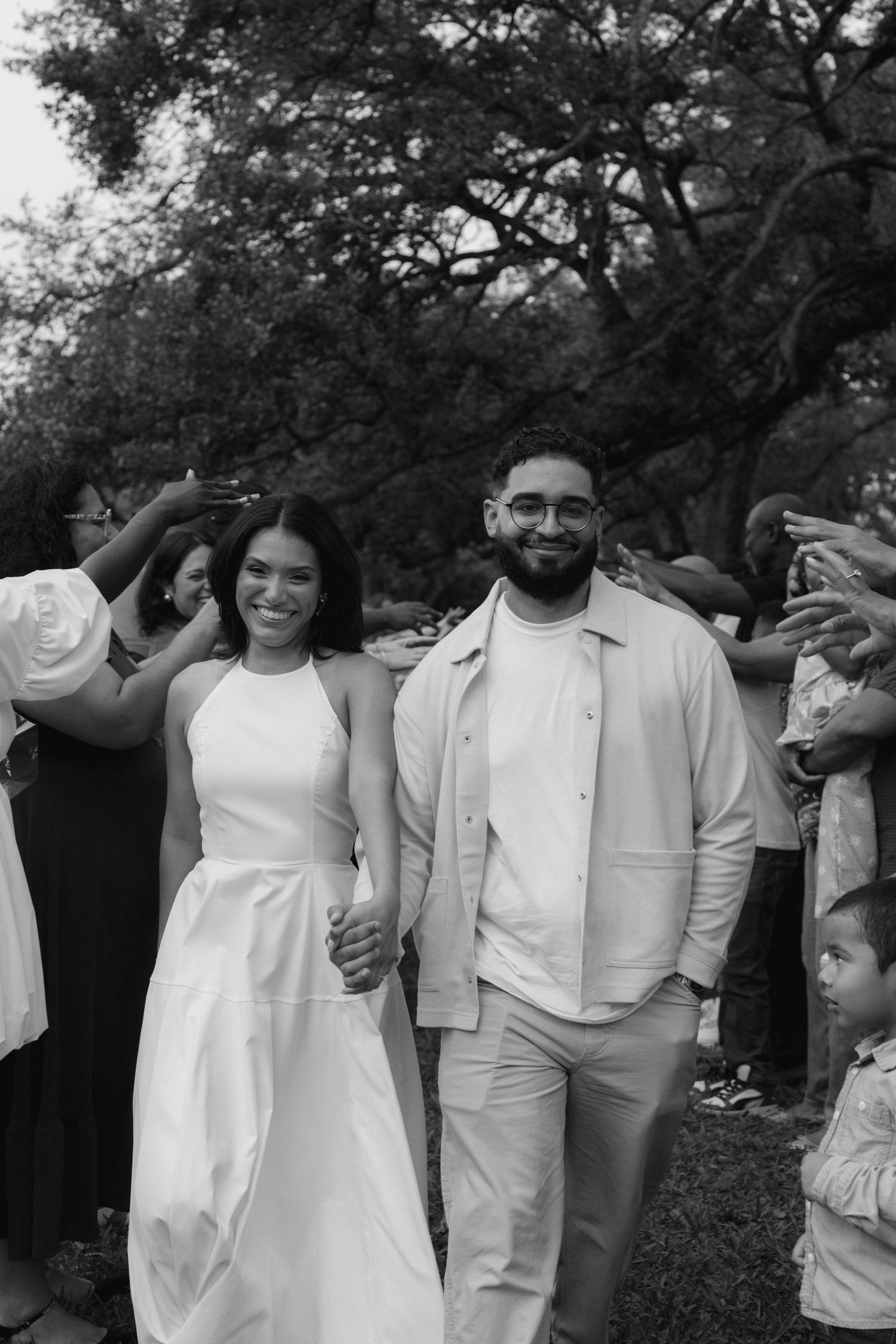Black and white photo of a smiling couple in wedding attire walking hand in hand outdoors among cheering guests.