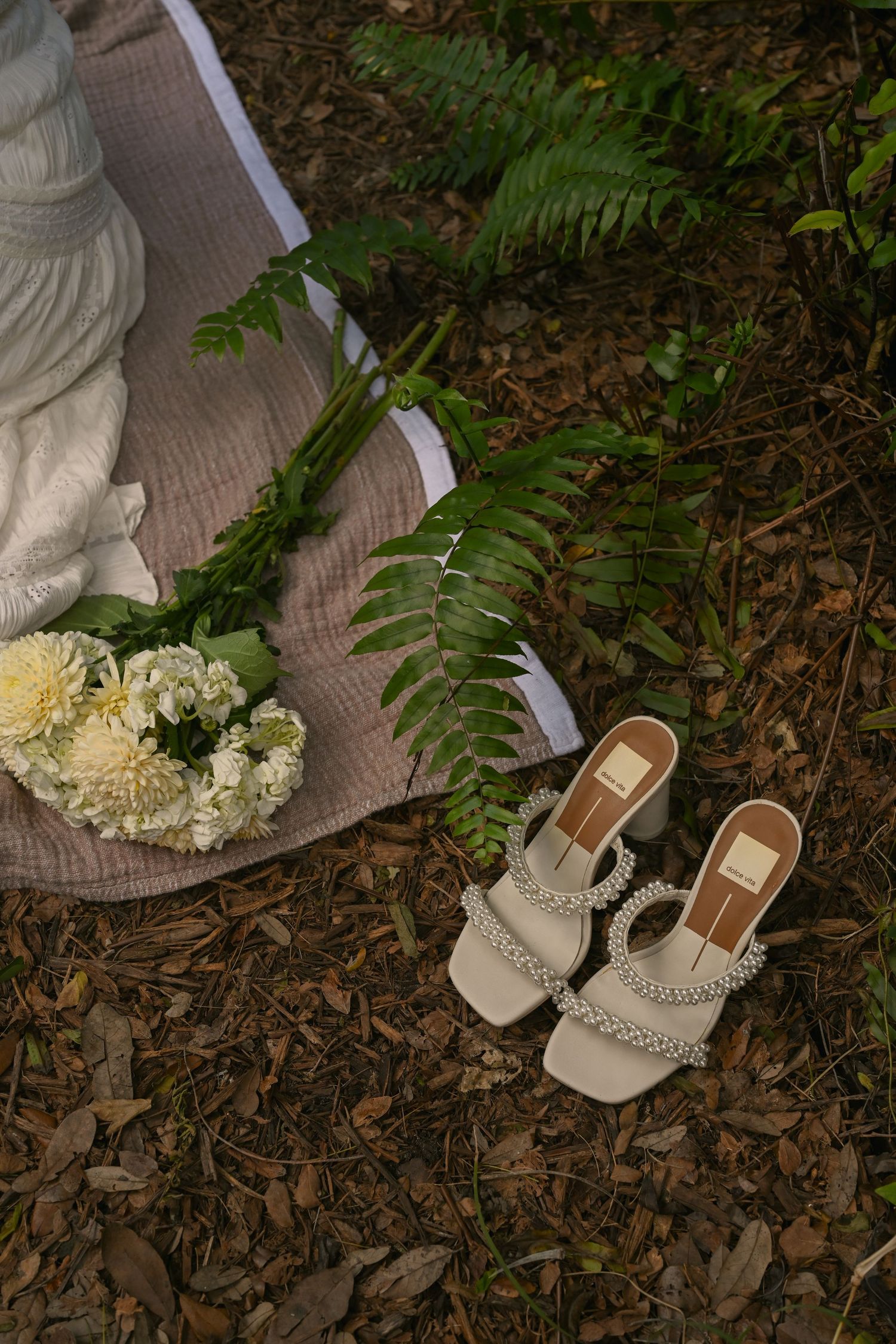 Elegant beige heeled sandals with pearl straps beside white flowers and fern leaves on woodland ground.