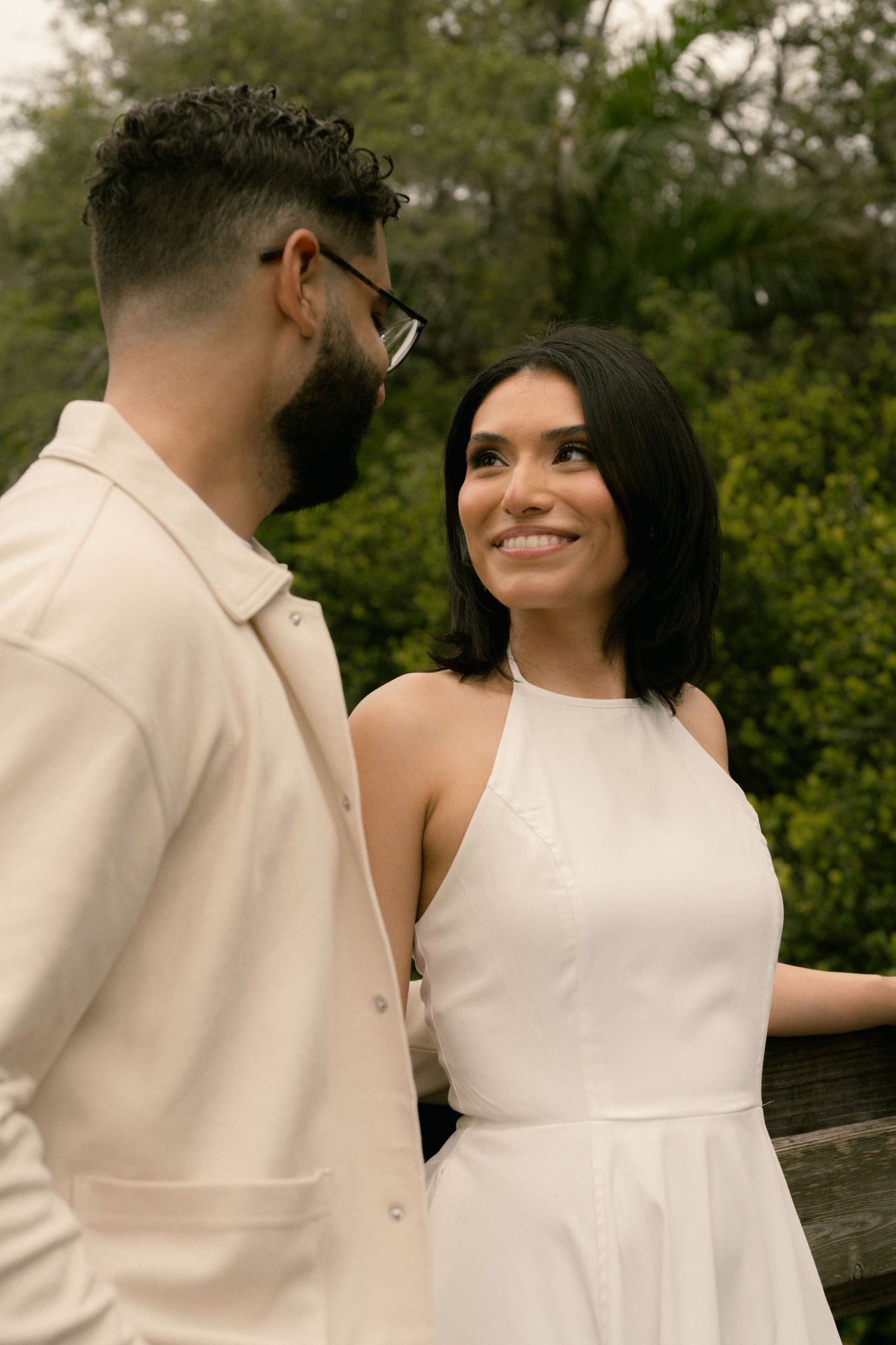 Smiling woman in white halter dress gazes at bearded man with glasses outdoors.