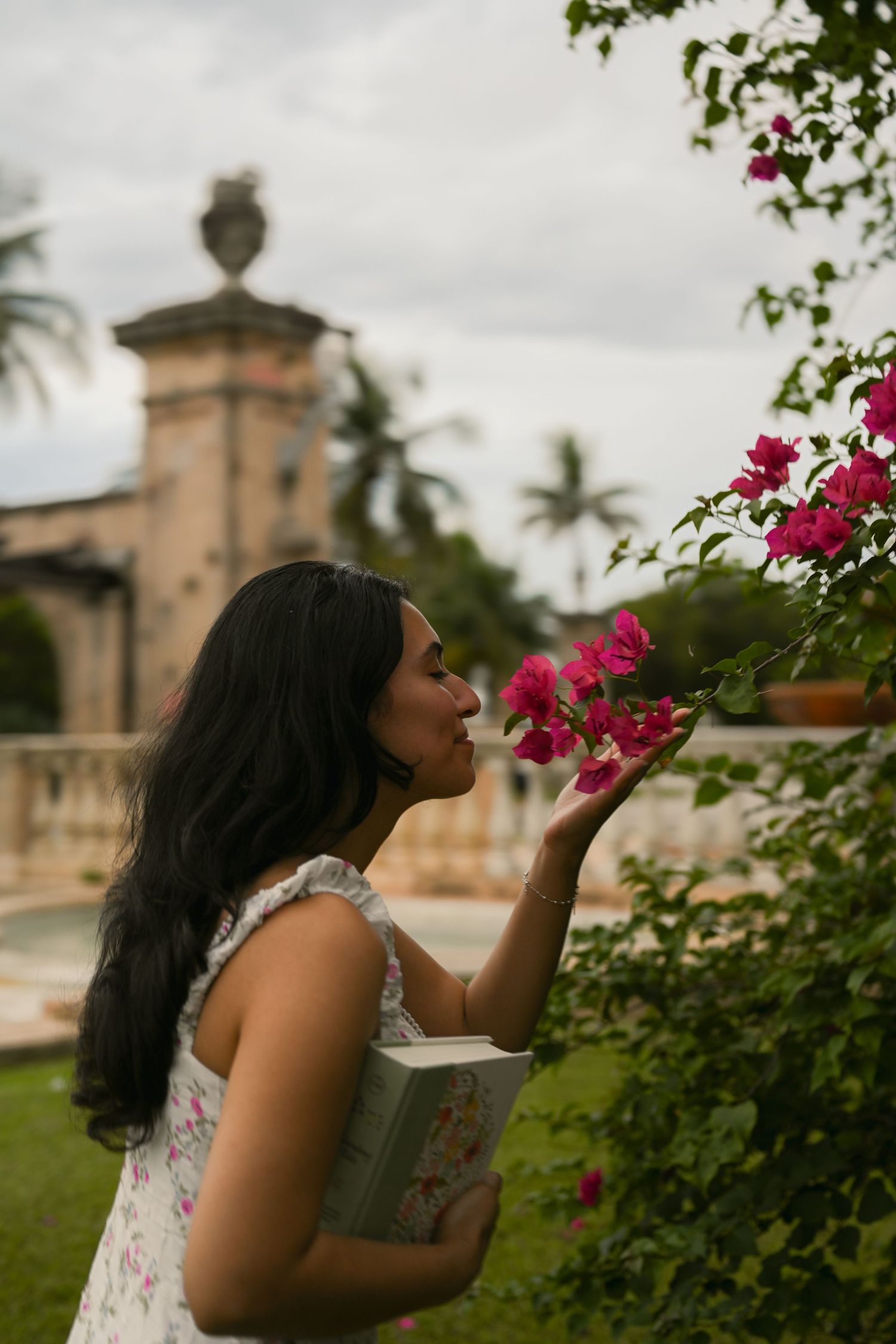 Woman in floral dress smelling pink bougainvillea flowers in a lush garden with historic architecture in background.