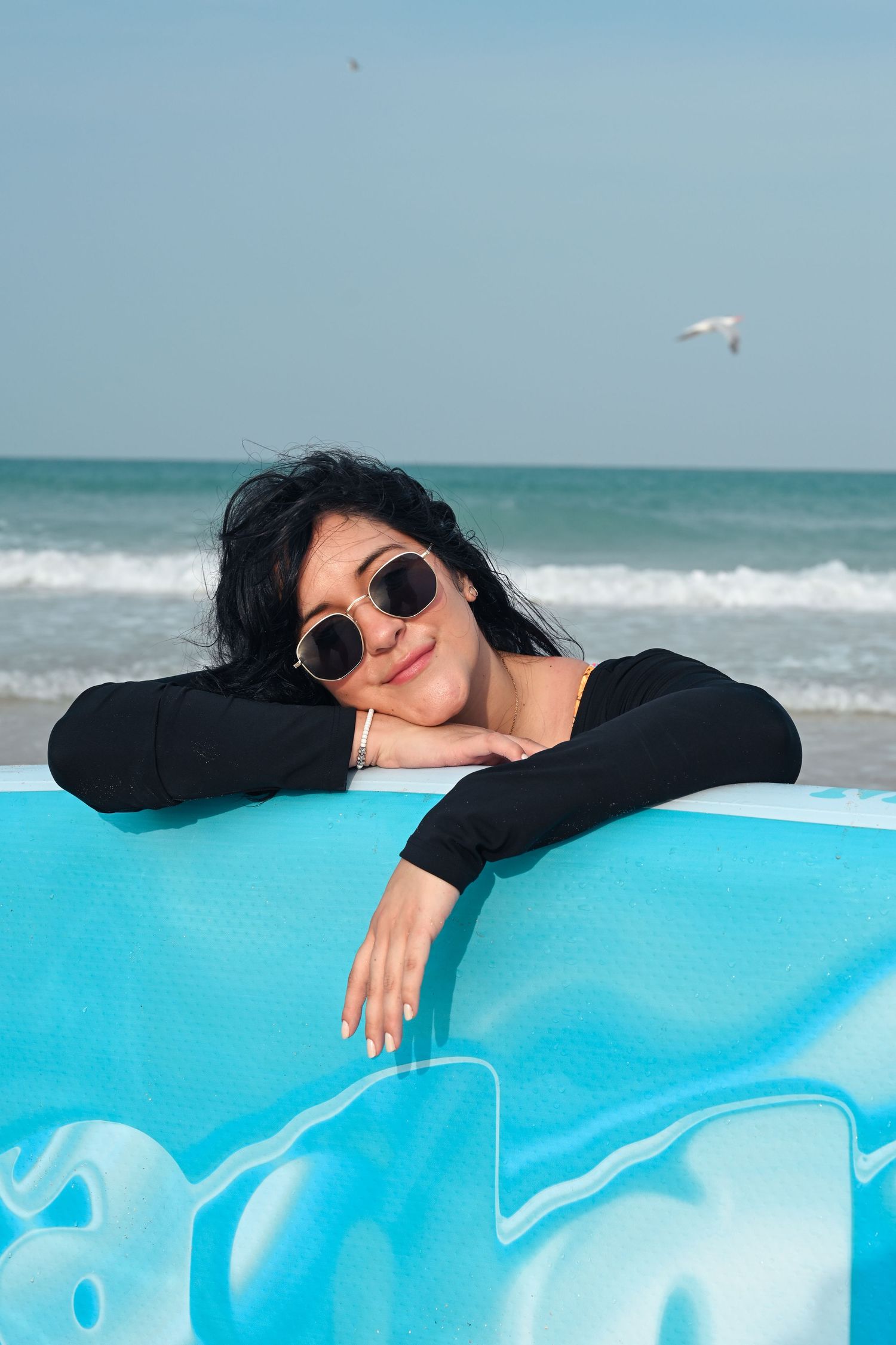 Person wearing sunglasses relaxes on turquoise surfboard by the ocean.