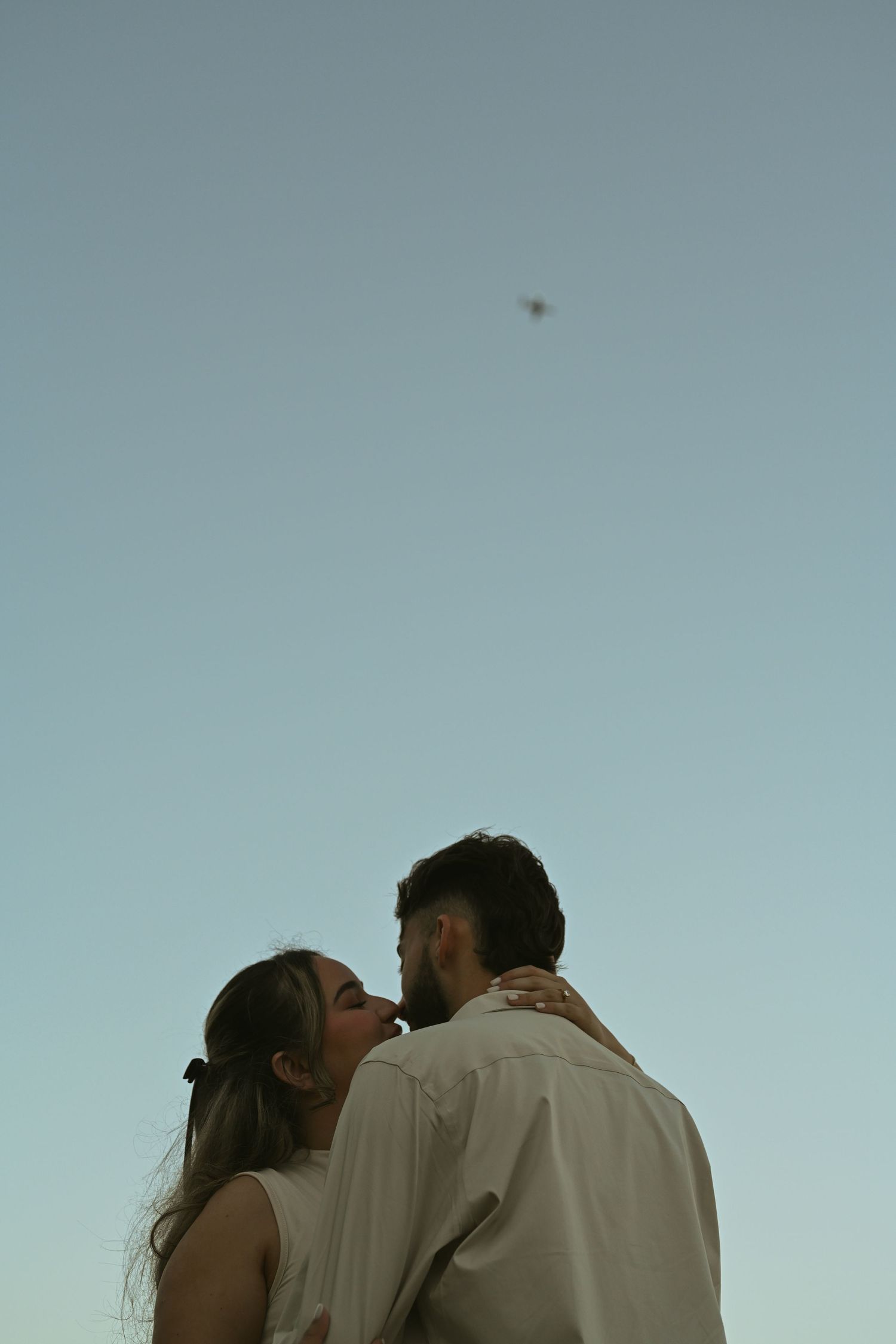 A romantic silhouette of a couple kissing against a pale blue sky at dusk.