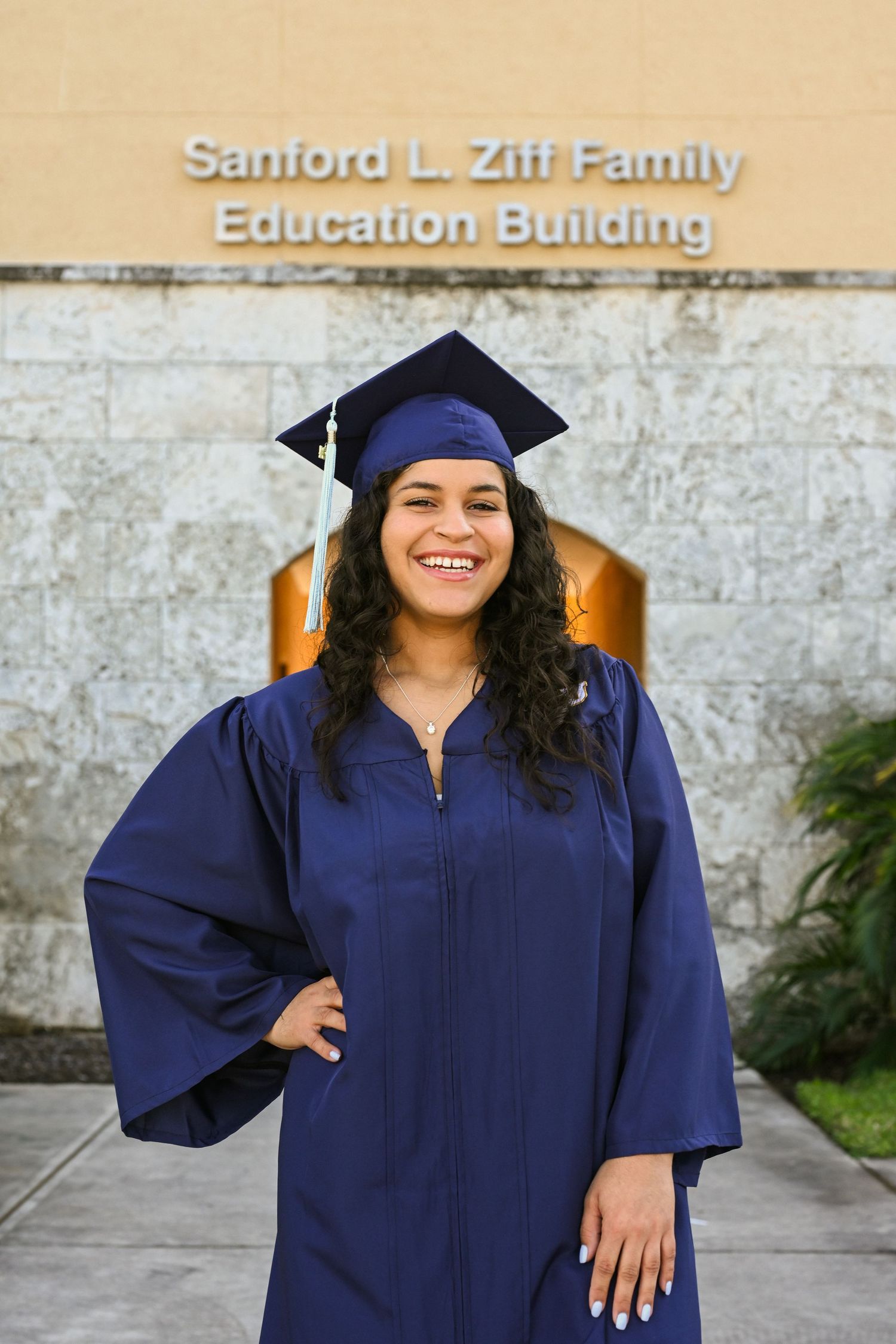 A graduate in navy blue cap and gown poses outside an education building.