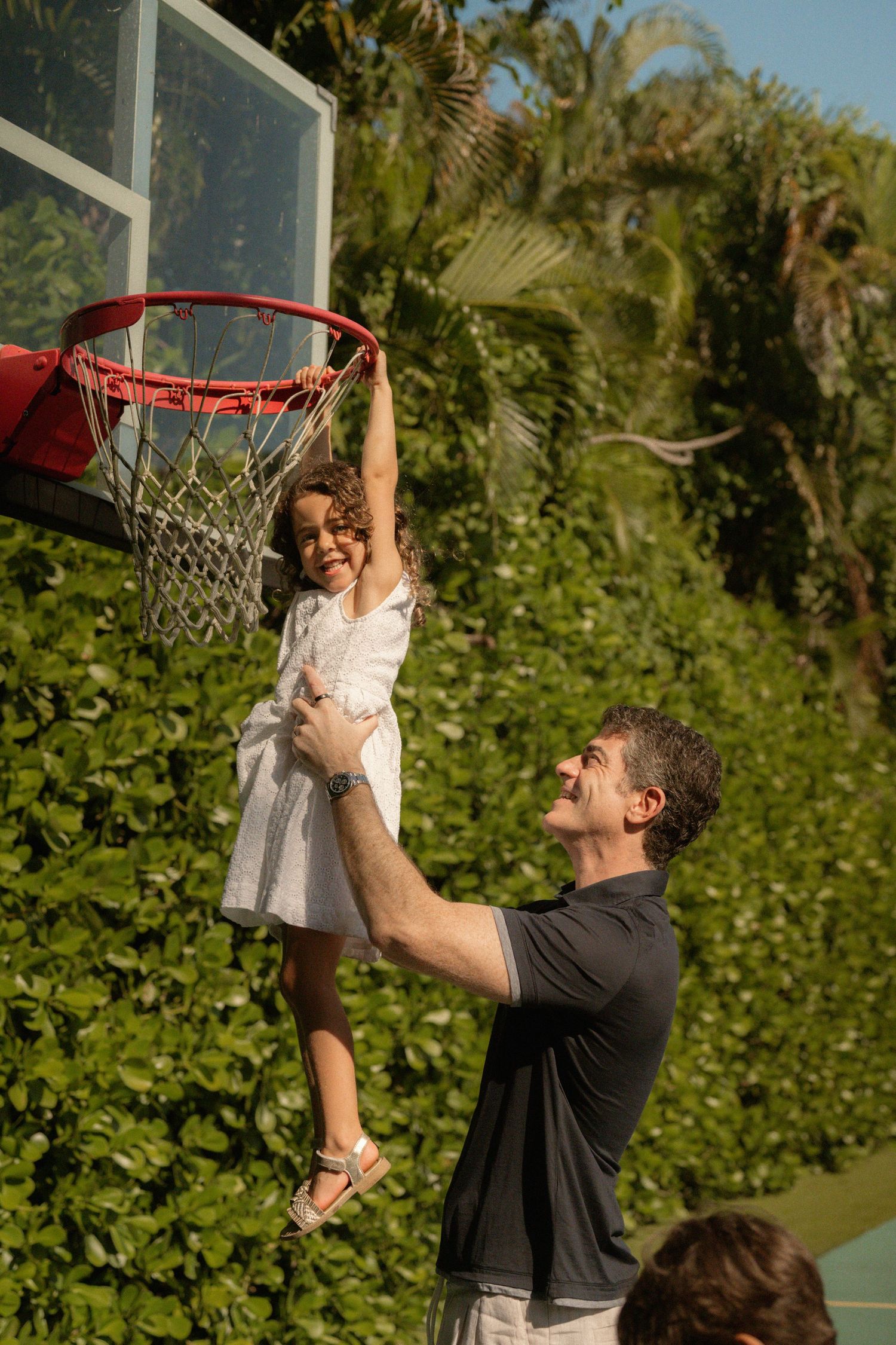 A man lifts a smiling young girl in a white dress so she can reach a basketball hoop, with lush green hedges behind them.