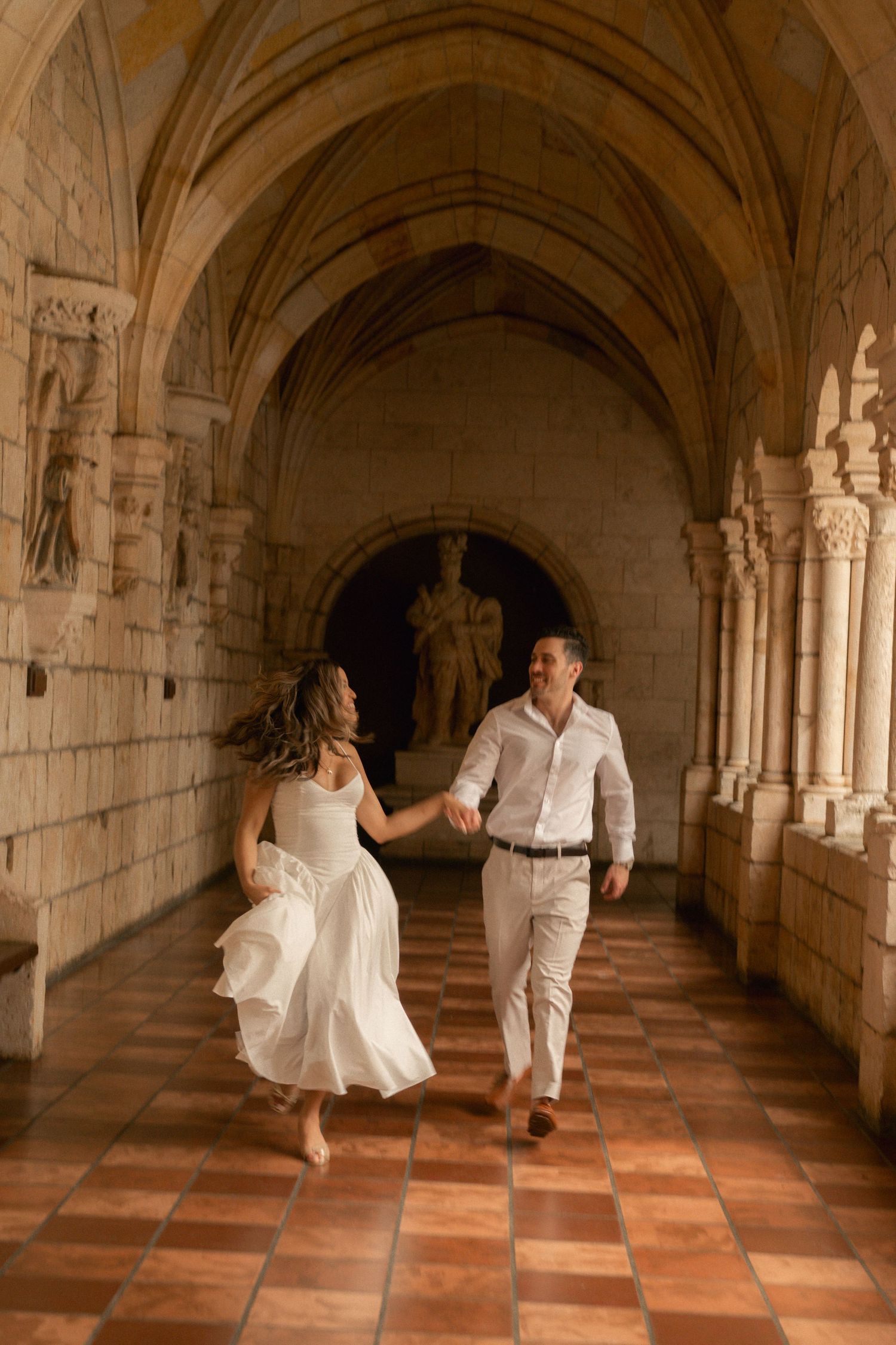 Couple in white outfits holding hands and running barefoot through a historic stone cloister with arched ceilings and terracotta floors.