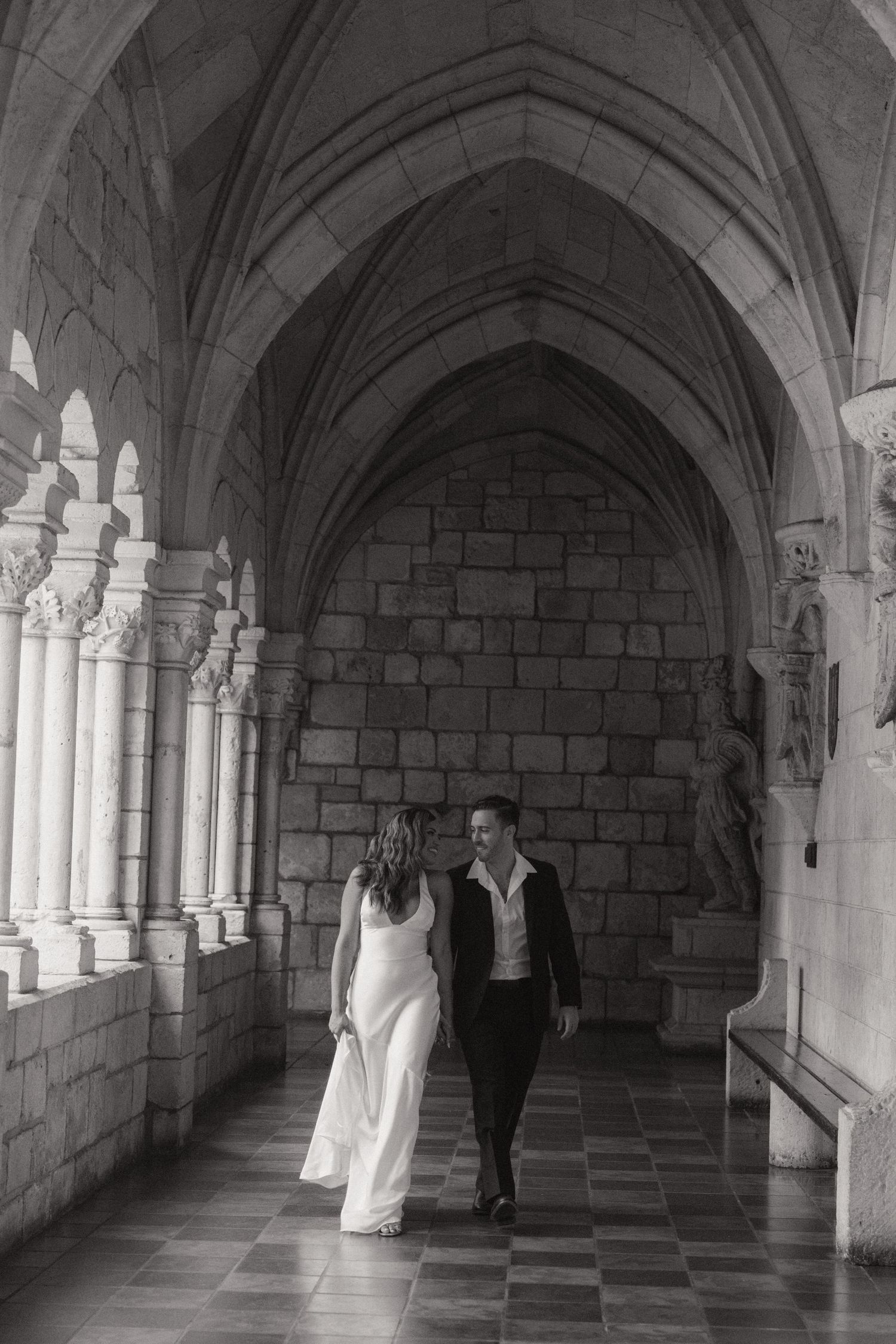 Black and white photo of a couple walking hand in hand through a historic stone cloister with Gothic arches and carved columns.