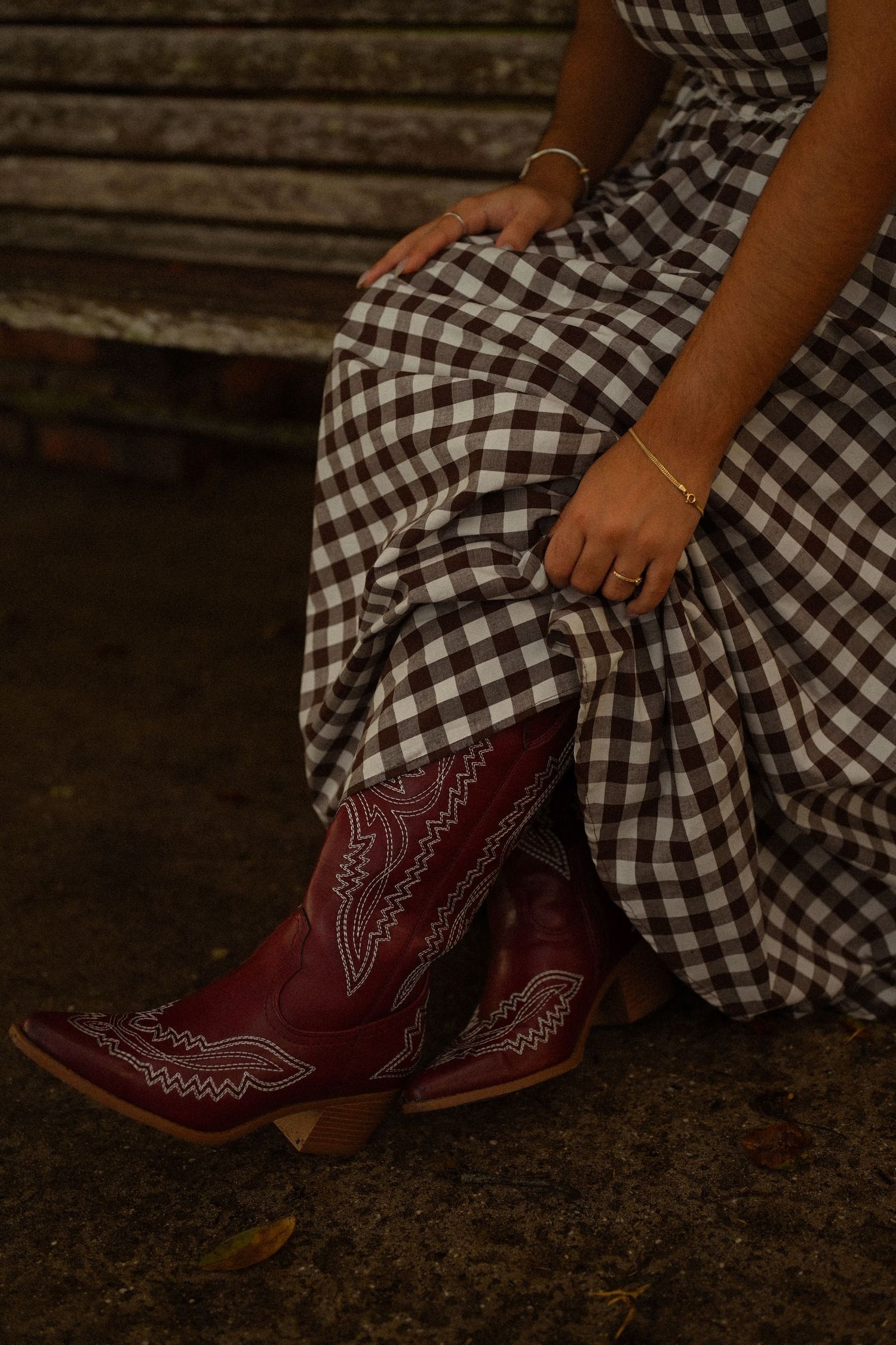 Red cowboy boots peek out from under a black and white checkered dress on a wooden bench.