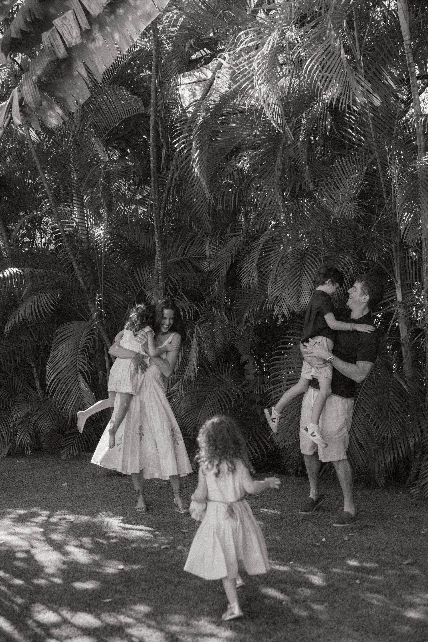 Black and white family photo of two parents lifting and playing with three children beneath tall tropical palm trees in dappled sunlight.