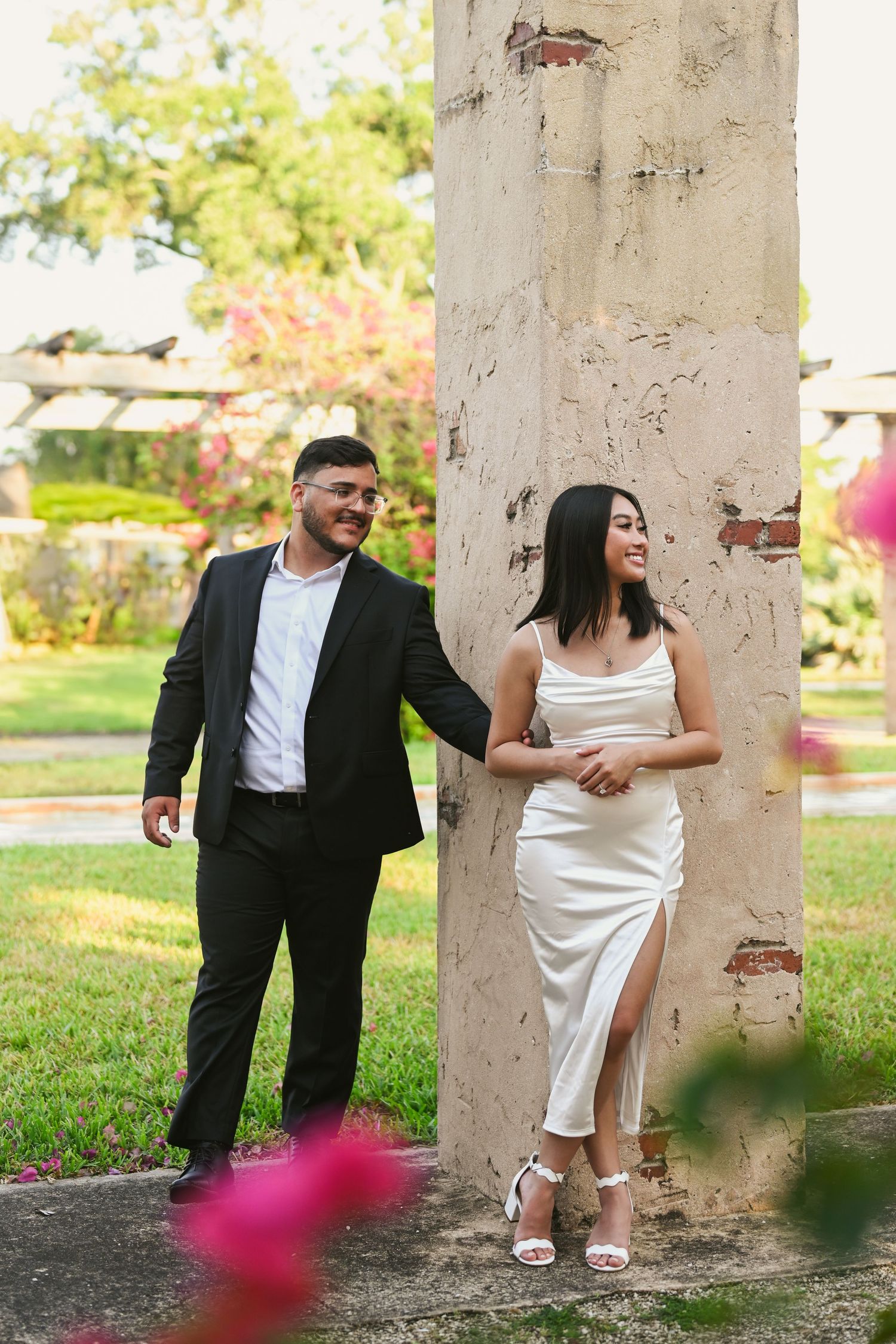 Elegant couple posing playfully between stone columns with pink flowers in the background.