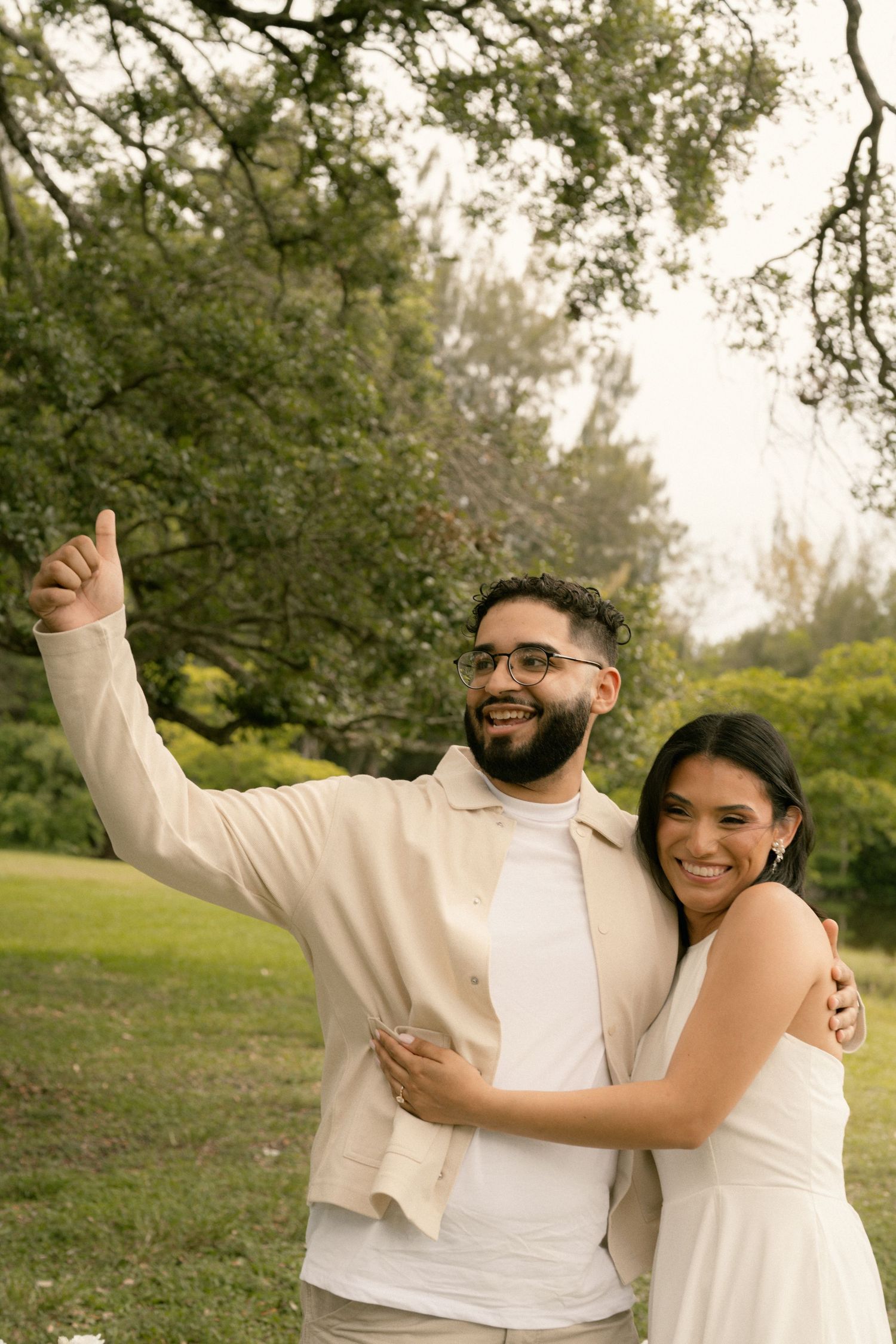 Smiling couple embraces outdoors, man pointing up wearing beige jacket, woman in white dress, lush green trees background.