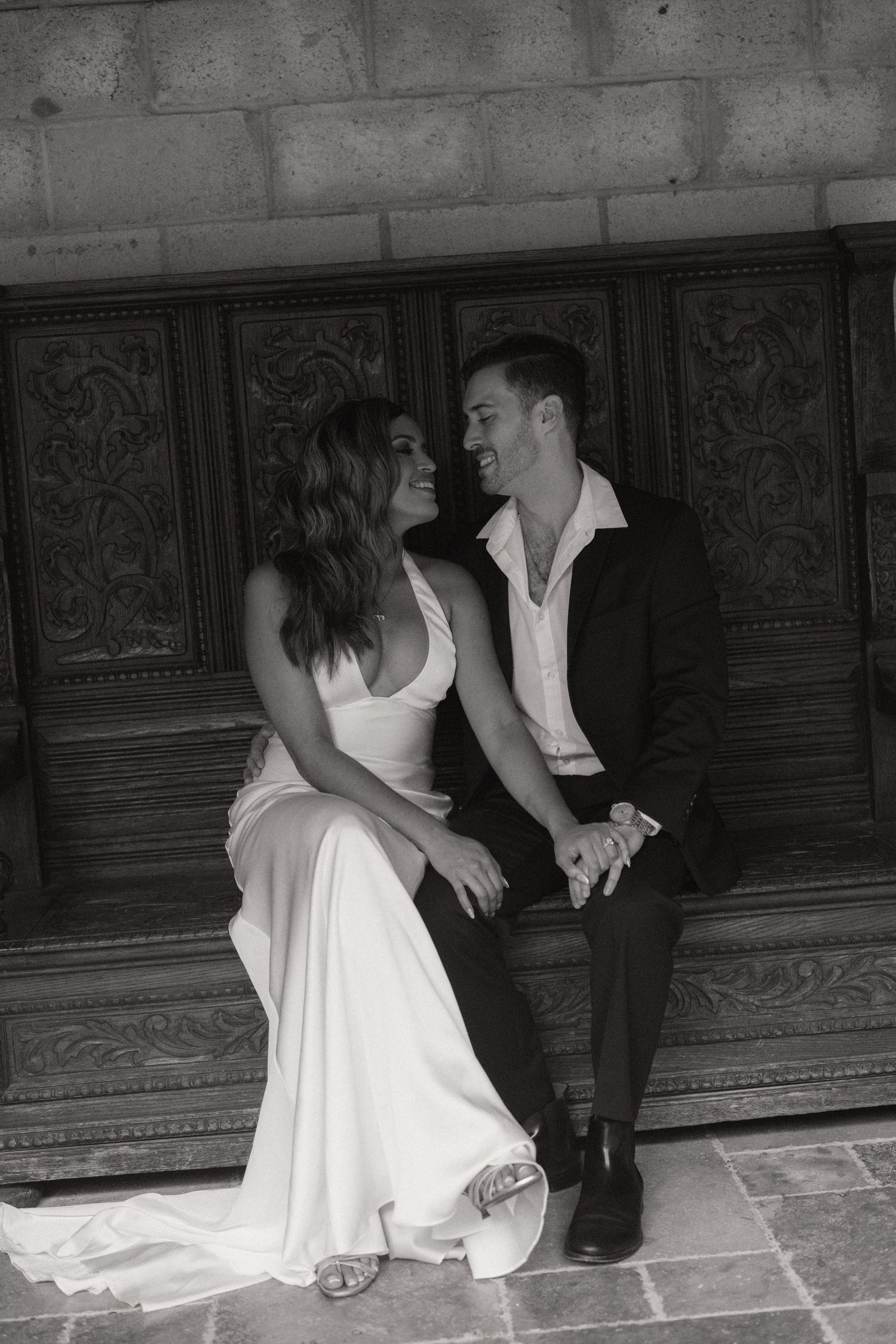 Black and white photo of a smiling couple in elegant formal attire seated on a carved wooden bench against a stone wall.