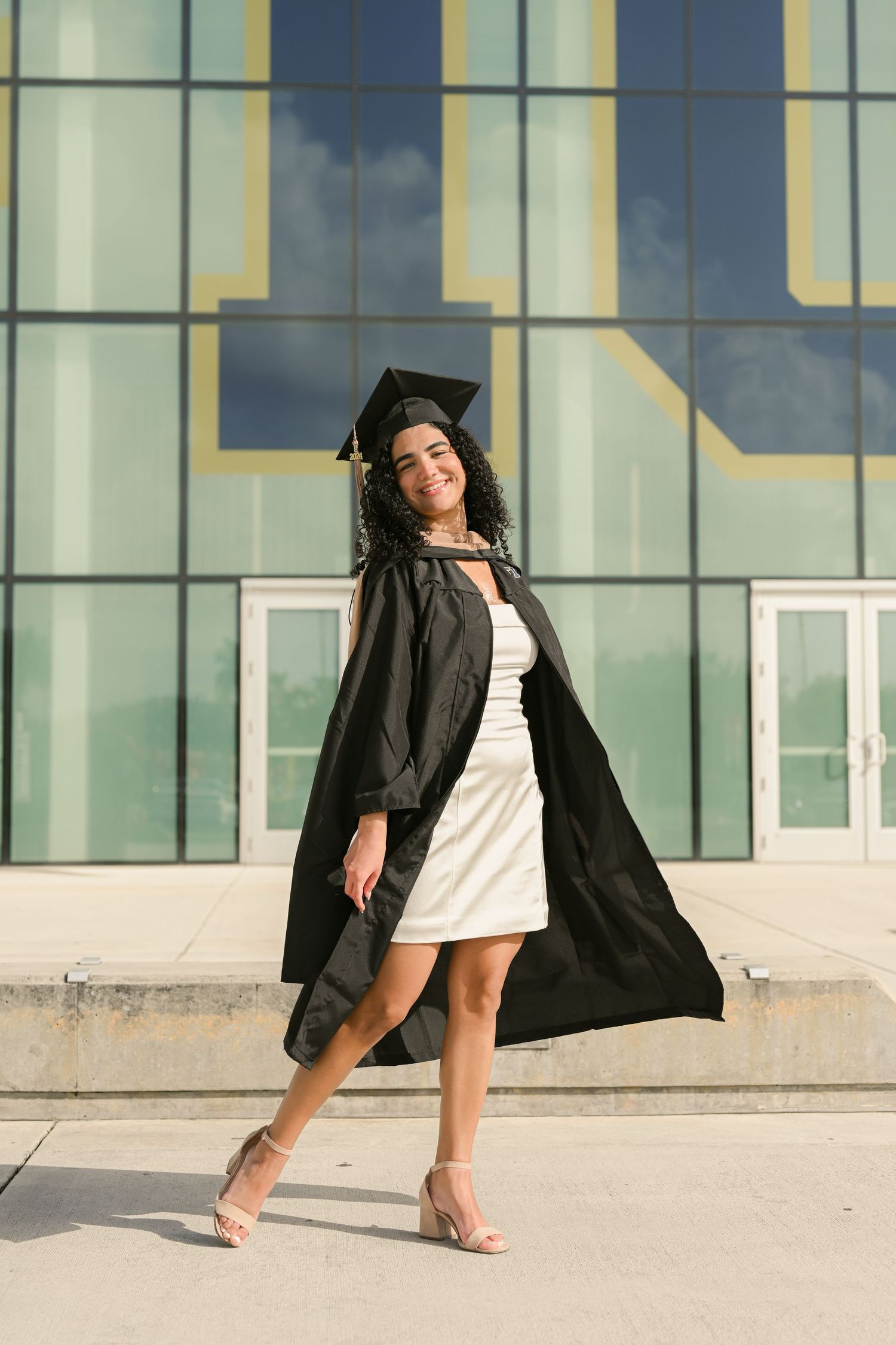 Graduate in black gown dances playfully on campus during graduation photoshoot.