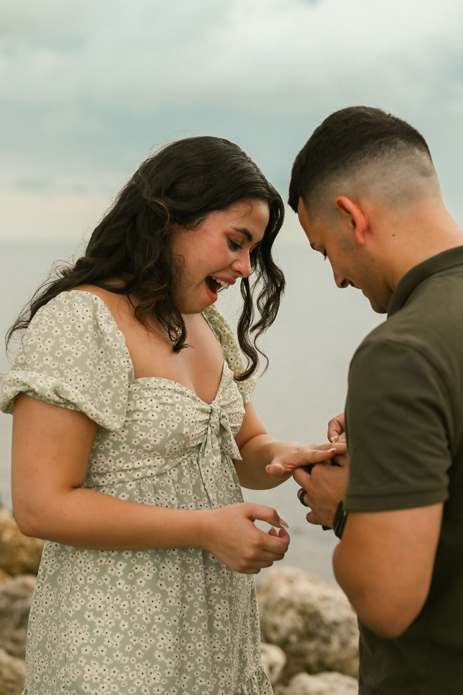 A couple examines an engagement ring during a seaside proposal.