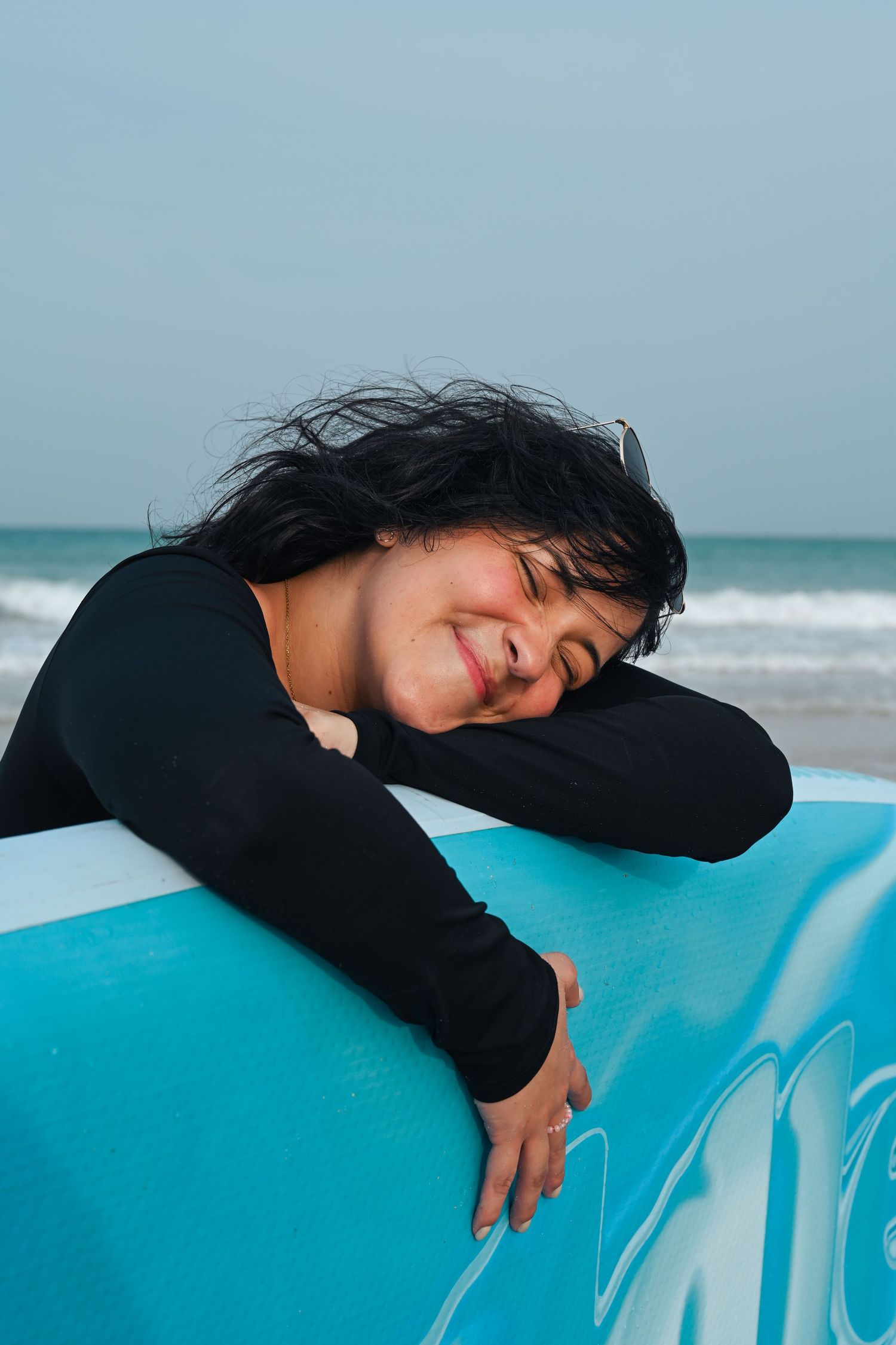 Person in black lounging on bright blue surfboard with ocean waves in background.