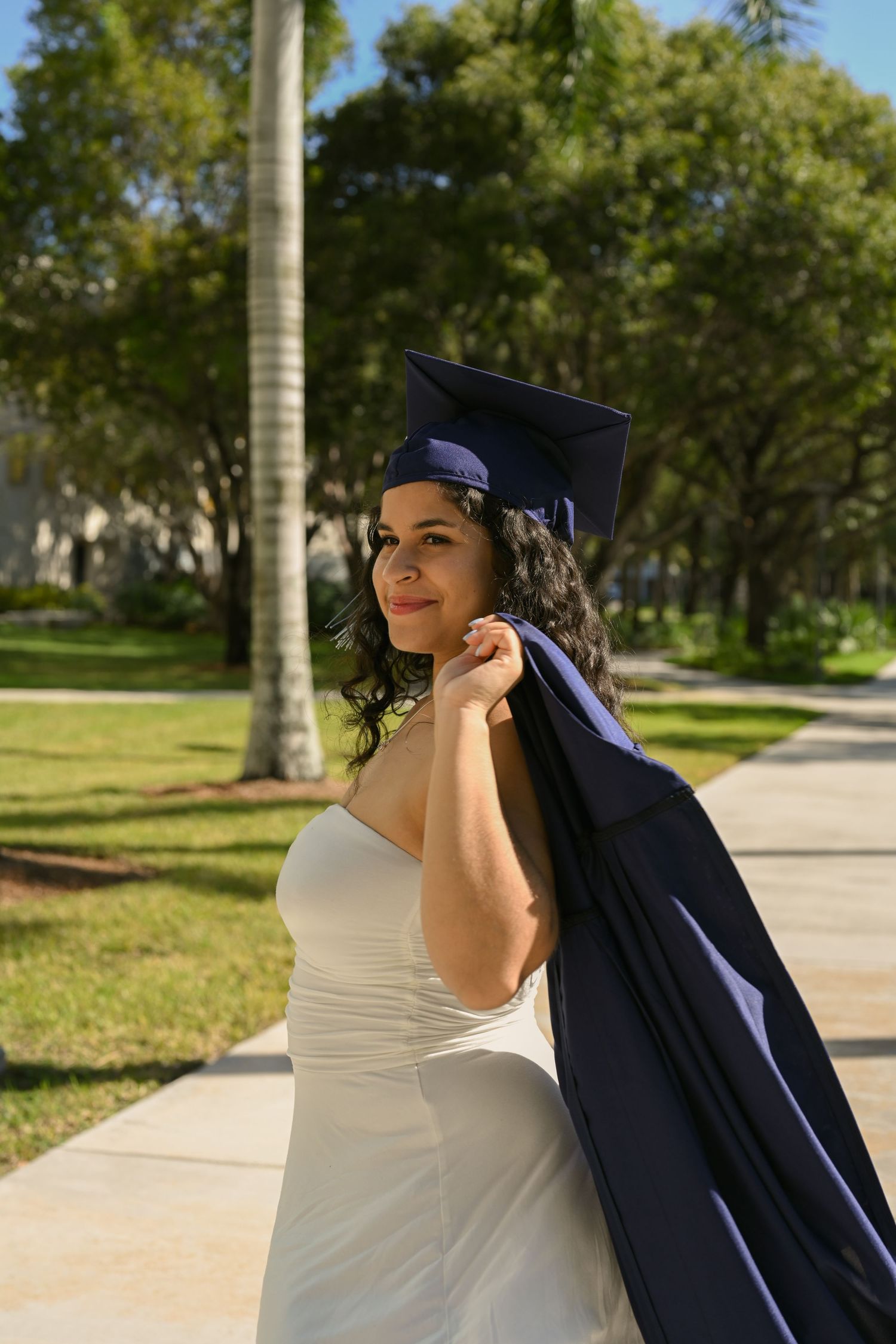 Graduate in white dress poses with graduation gown against palm trees on campus.